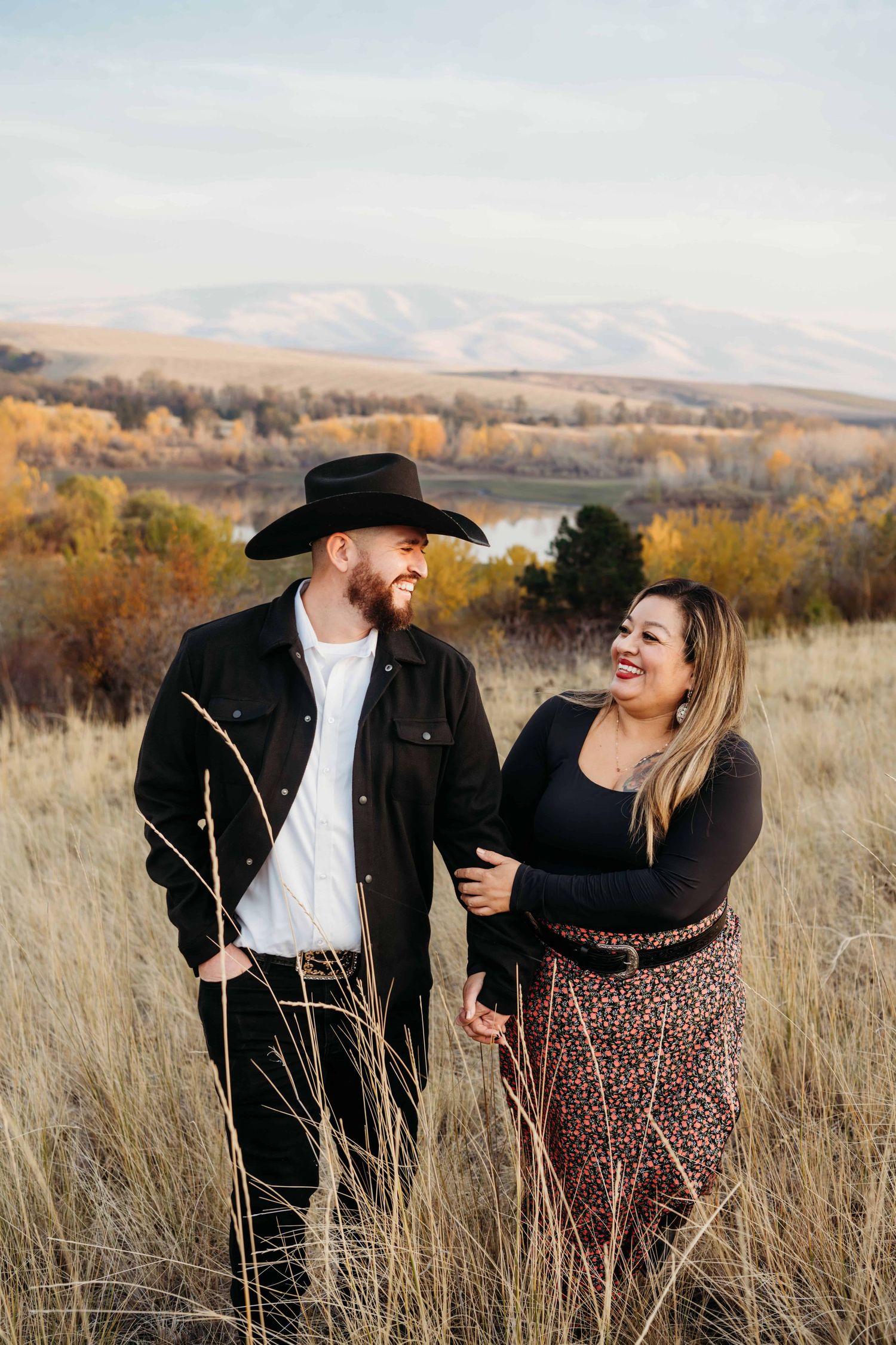 A man wearing a cowboy hat and his girlfriend walking through a field with a lake and hills in the background.