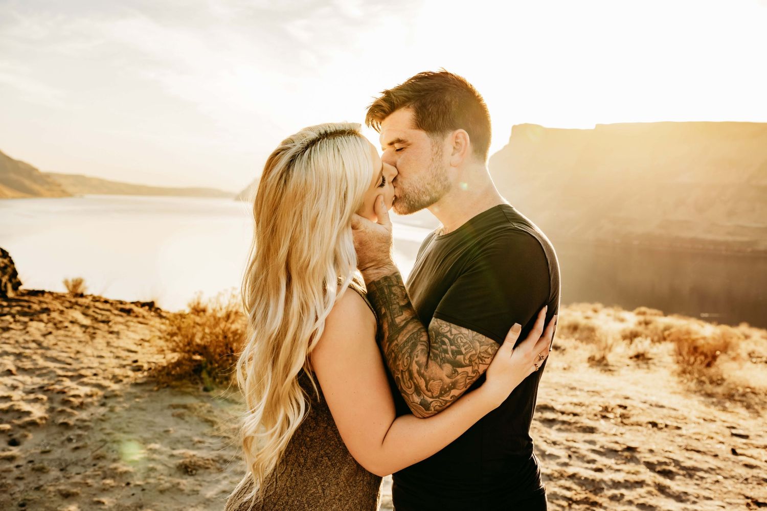 A portrait filled with golden lighting of a couple kissing in a sandy scene with a river and mountains in the background.