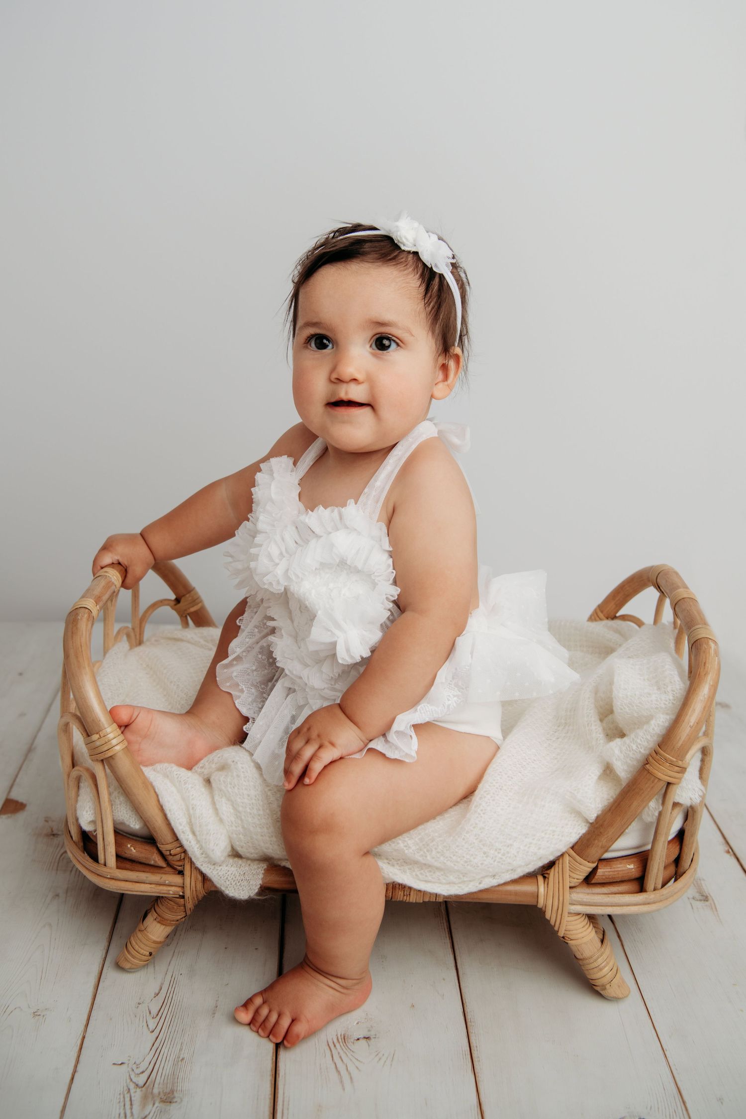A baby dressed in white sitting on a brown bed.
