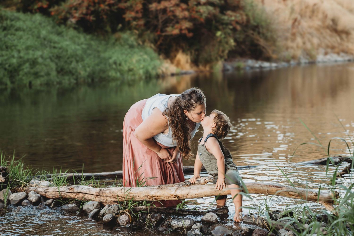A child sitting on a log giving his mother a kiss while they play together in a creek.