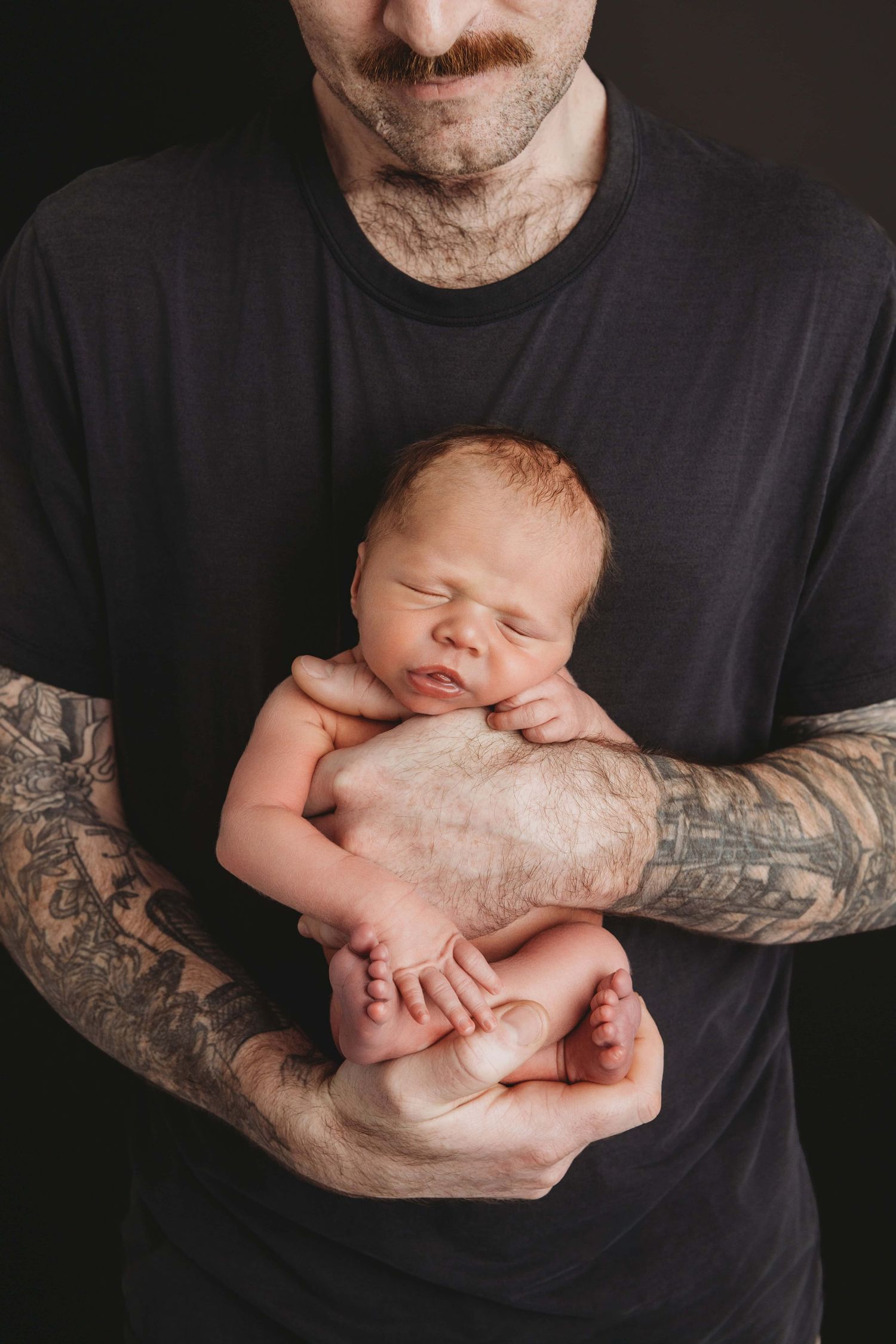 A high contrast portrait of a newborn in his tattooed dad's arms on a black backdrop.