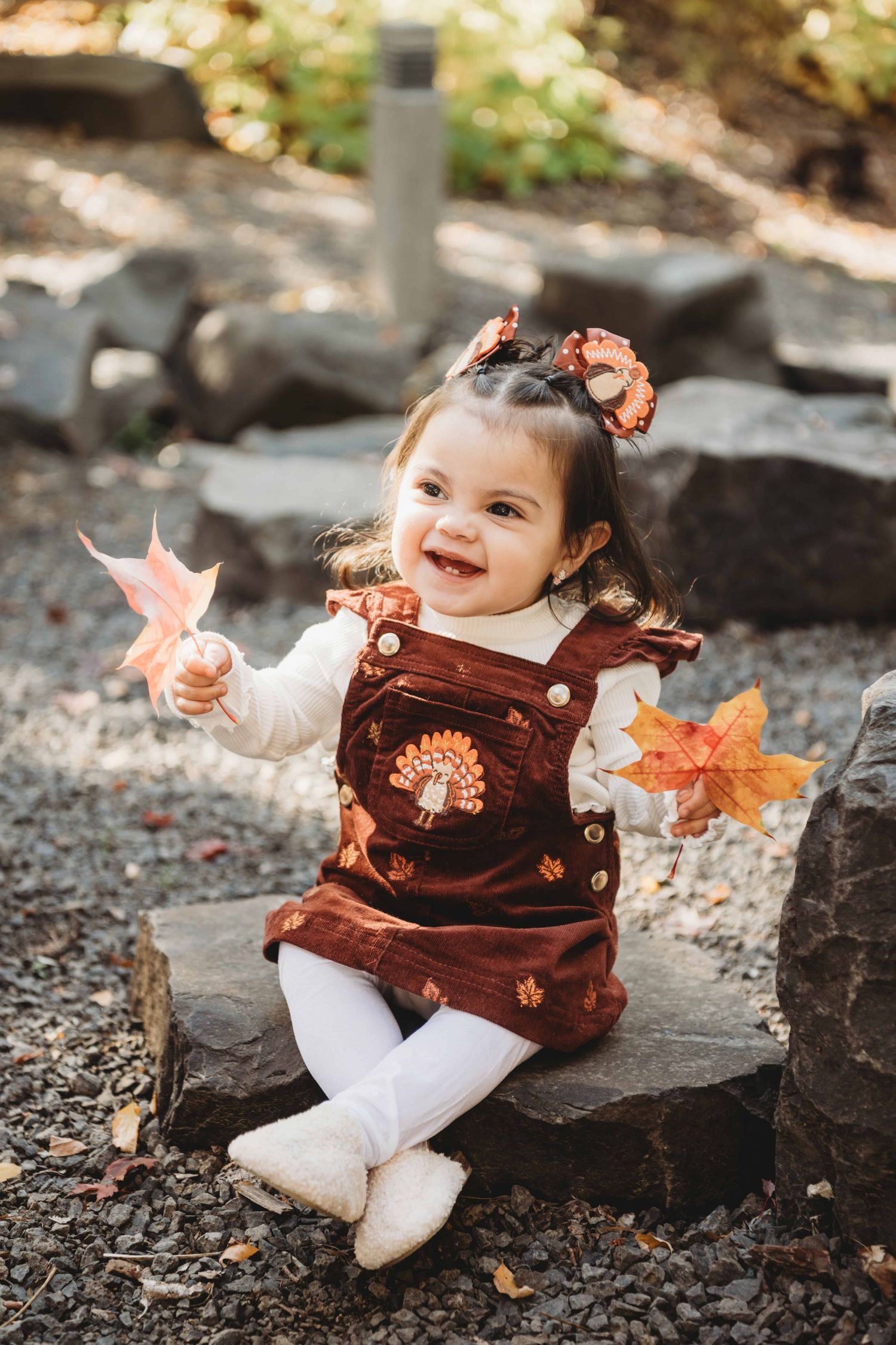 A baby girl sitting on a low rock, holding leaves and laughing.