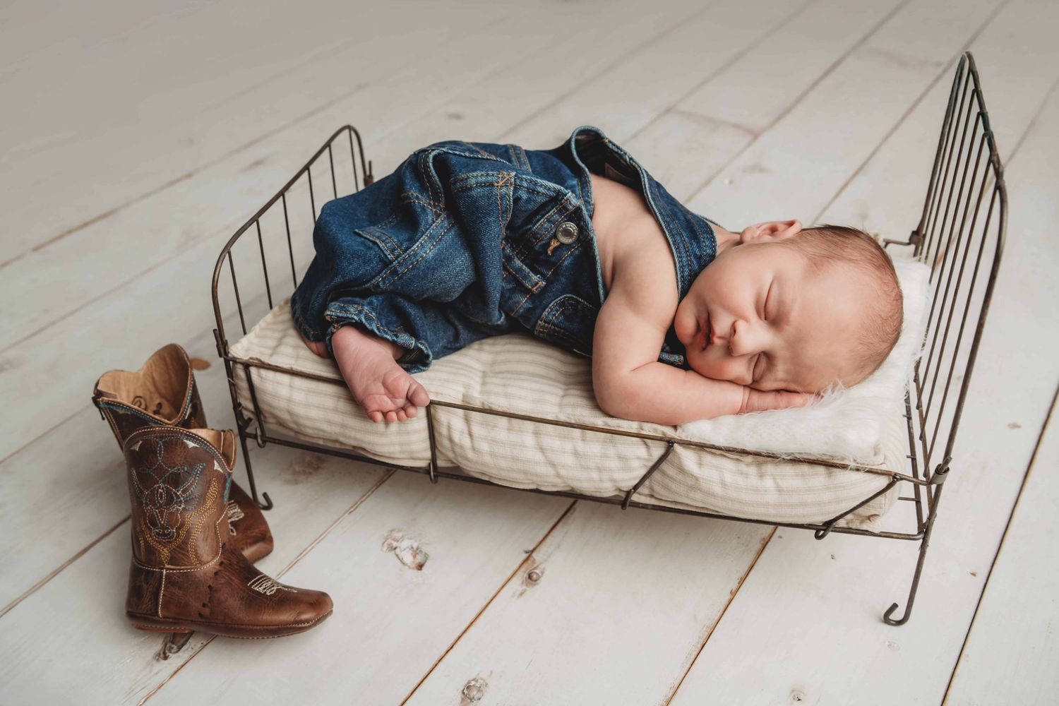 Studio photo of a newborn lying on a wire bed with cowboy boots propped against the bed.