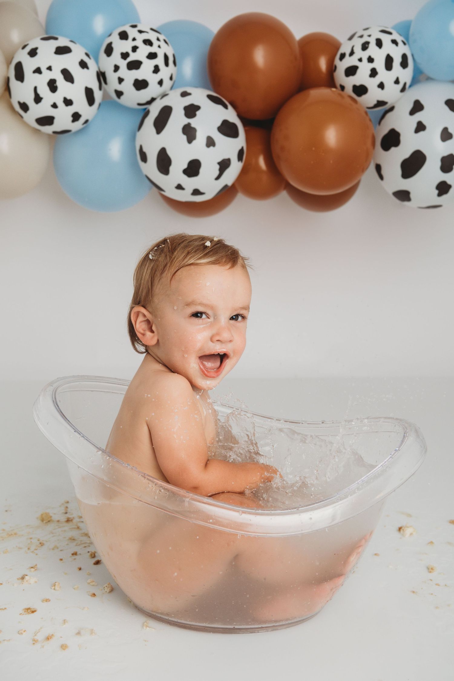 A one year old sitting in a clear bathtub with rodeo themed balloons in the background.