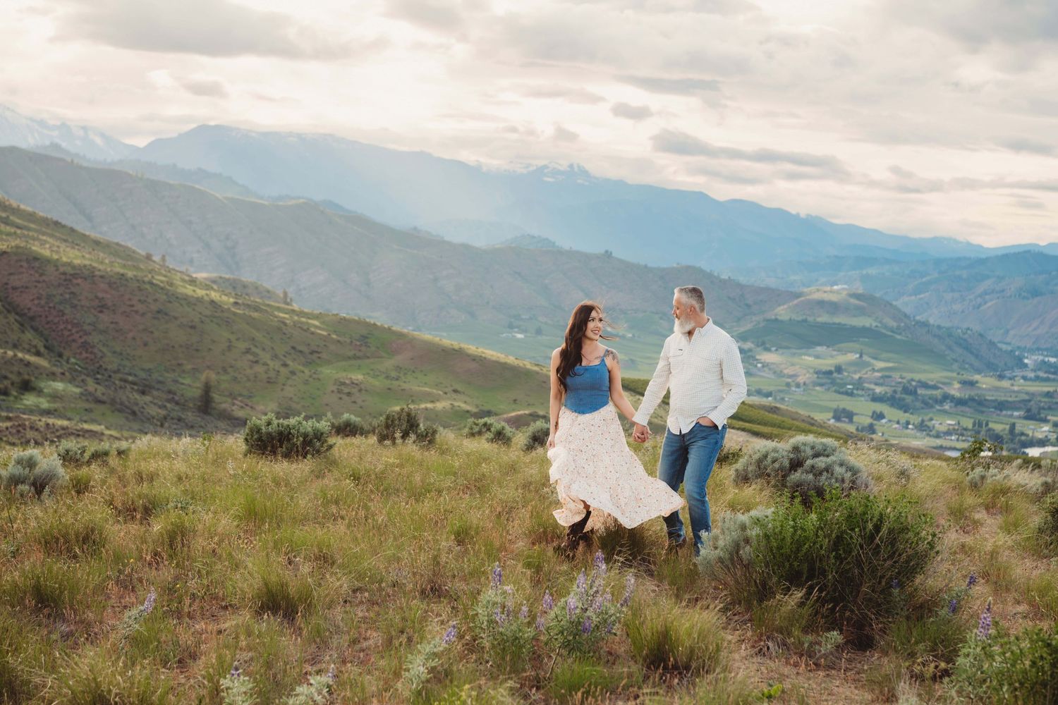 A portrait of a woman wearing a flowy dress holding hands with her husband while walking through a colorful field.