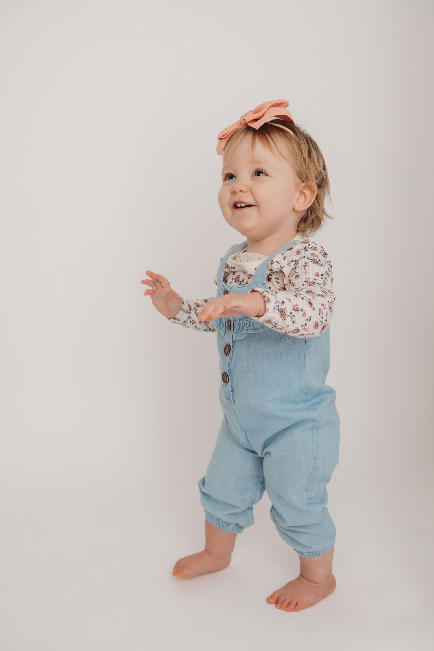A one year old wearing overalls and a pink bow walking on a white backdrop in studio