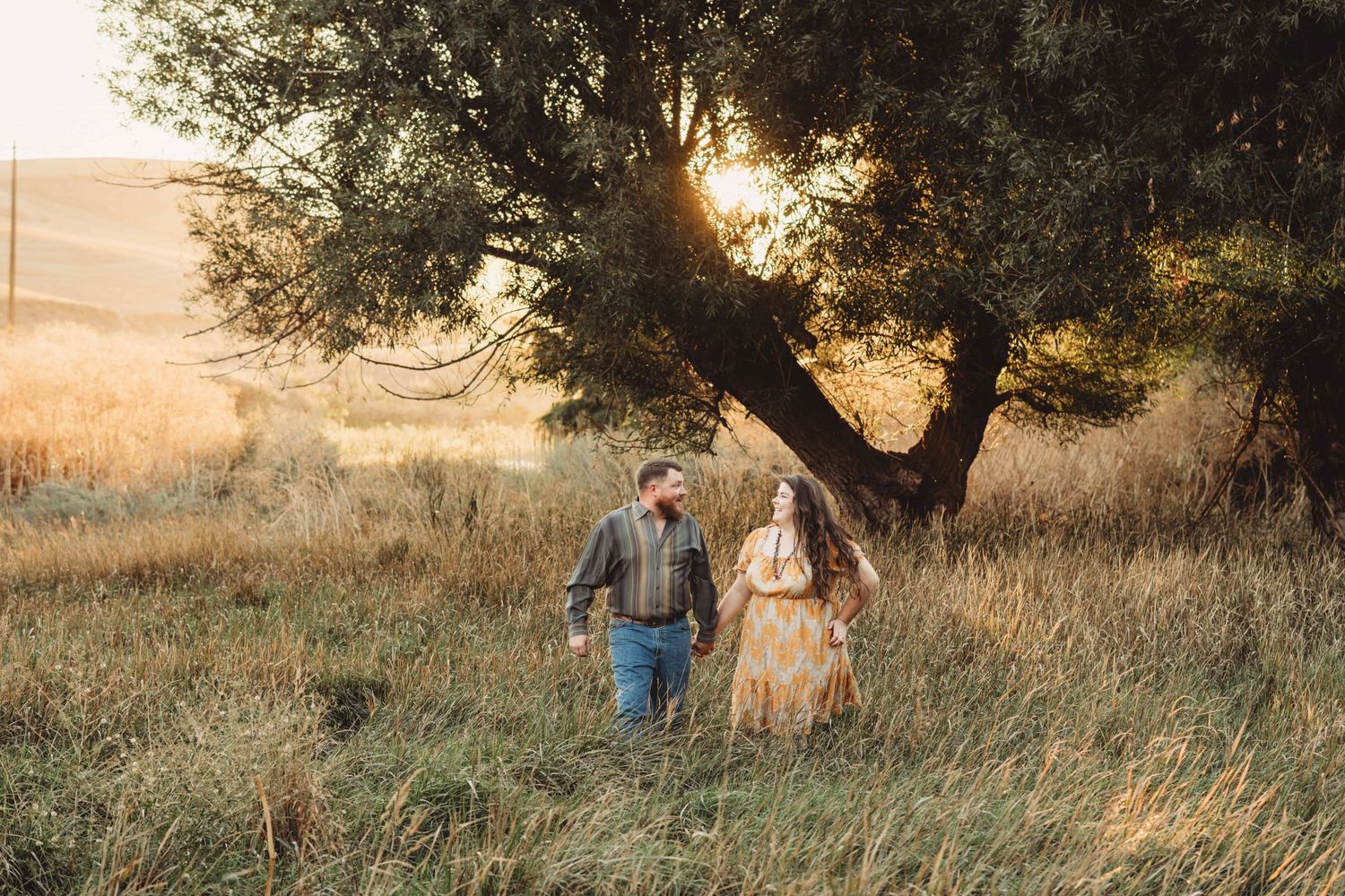 A couple walking through a field holding hands and looking at each other.