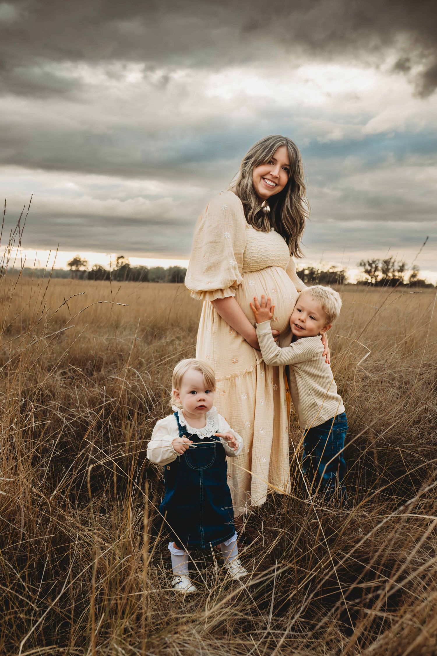 A boy hugging his mom's pregnant belly while his sister looks at the camera with a stormy sky.