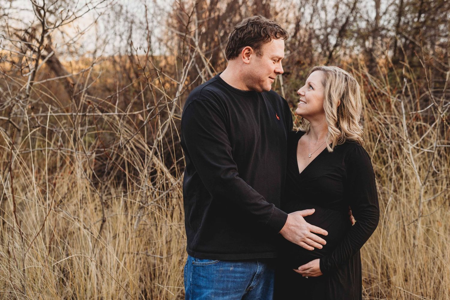 Outdoor maternity photography in Walla Walla showing an expectant couple embracing in tall grass with hands resting on her baby bump.