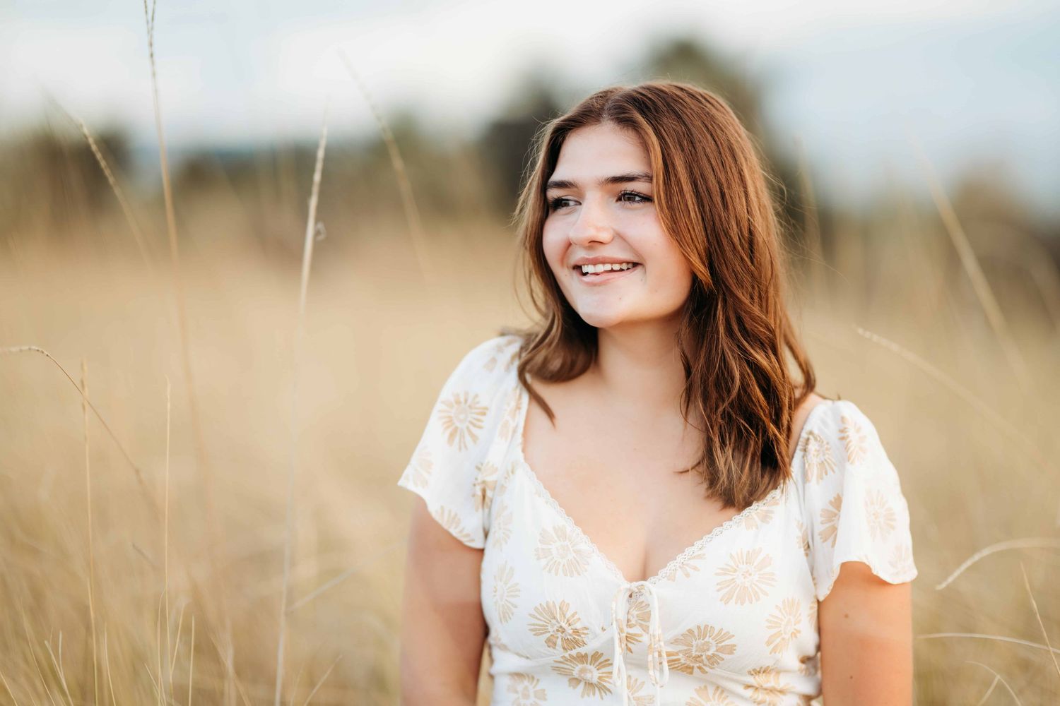 A senior portrait of a girl sitting surrounded by tall neutral grass and smiling.