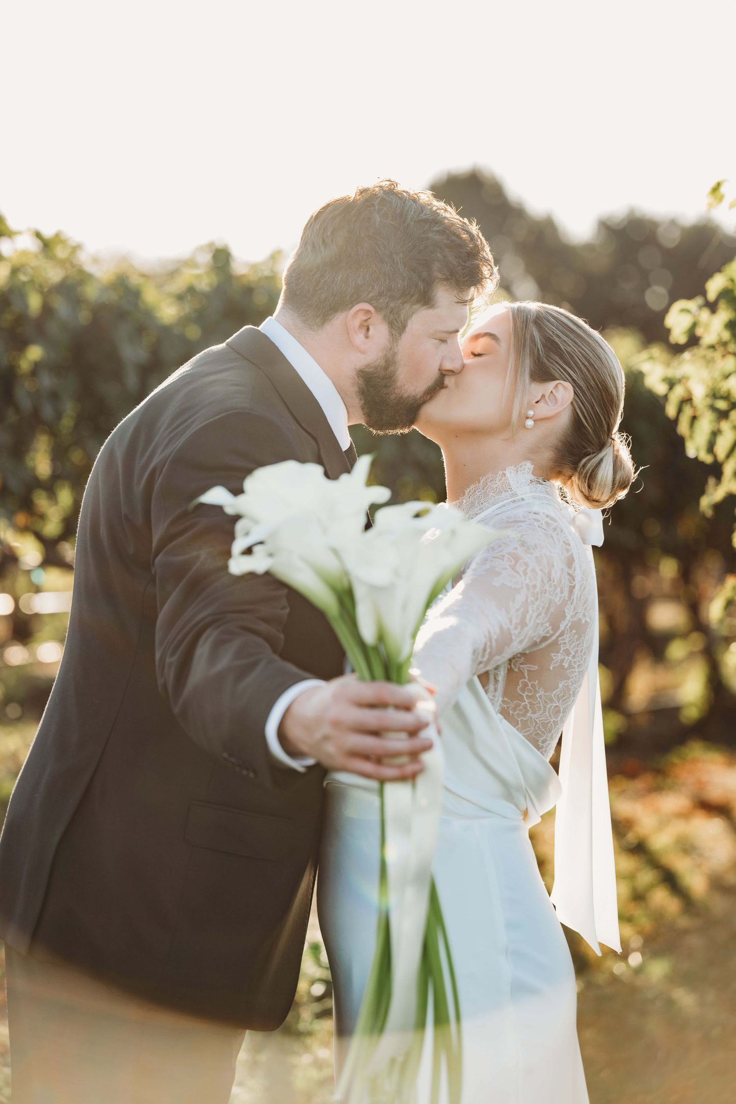 A bride and groom kissing and holding out their callalily bouquet on a sunny day.