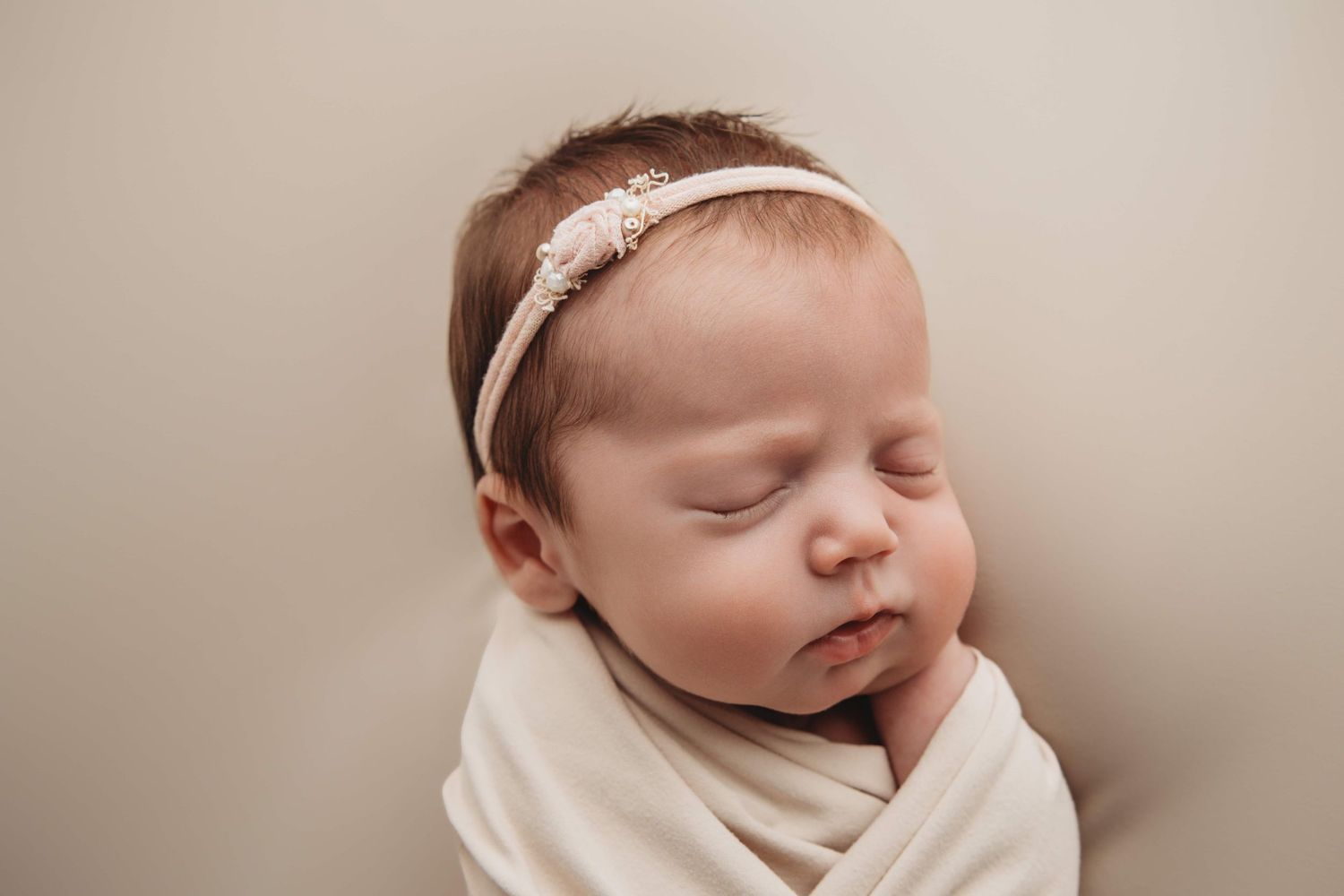 A newborn baby girl with chubby cheeks sleeping on a tan backdrop blanket with a simple dainty headband.