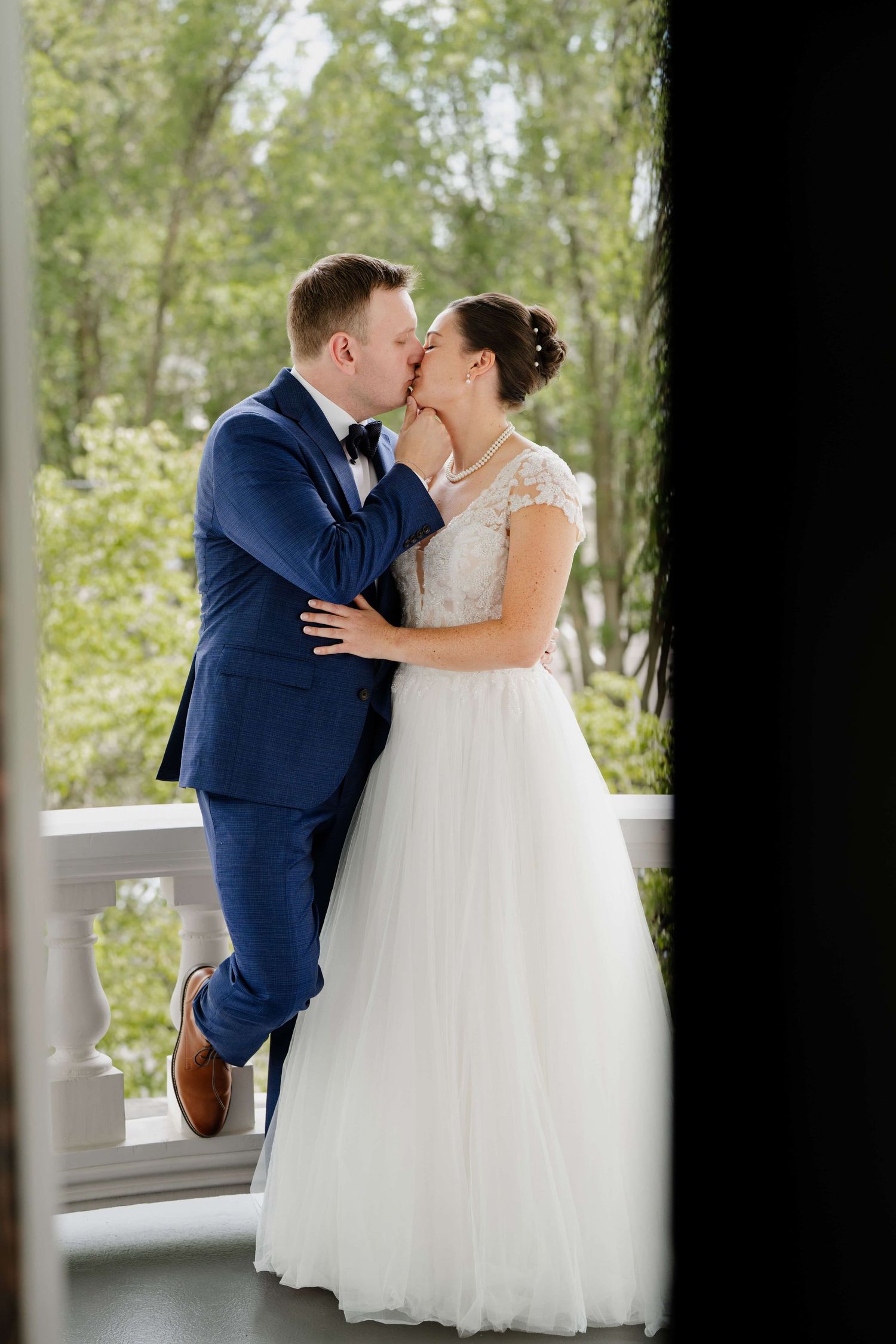 A couple in wedding attire kissing on a balcony, shot through a dark door frame.