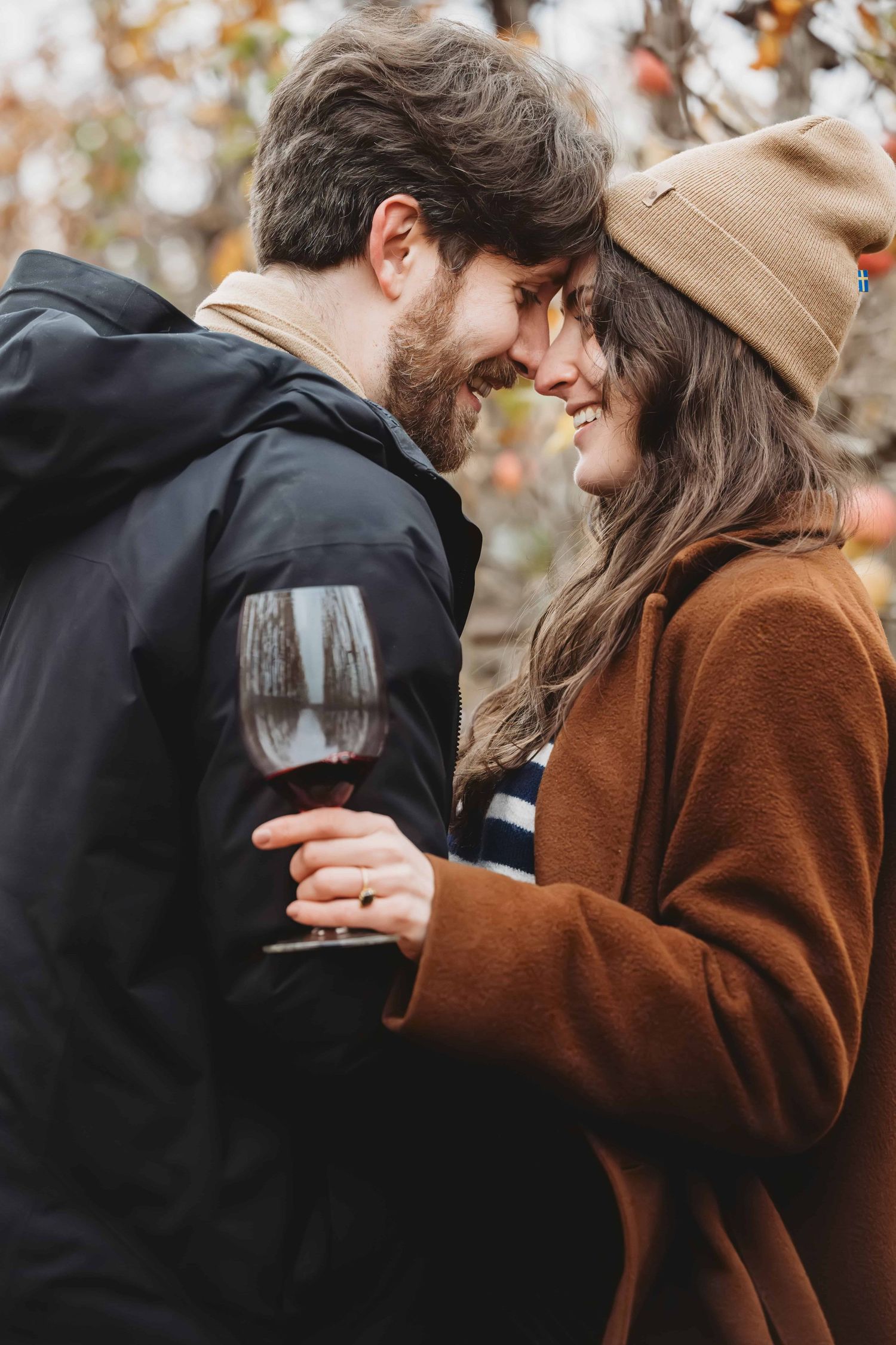 A couple standing in an orchard holding a wine glass, showing off her ring that he just proposed with.