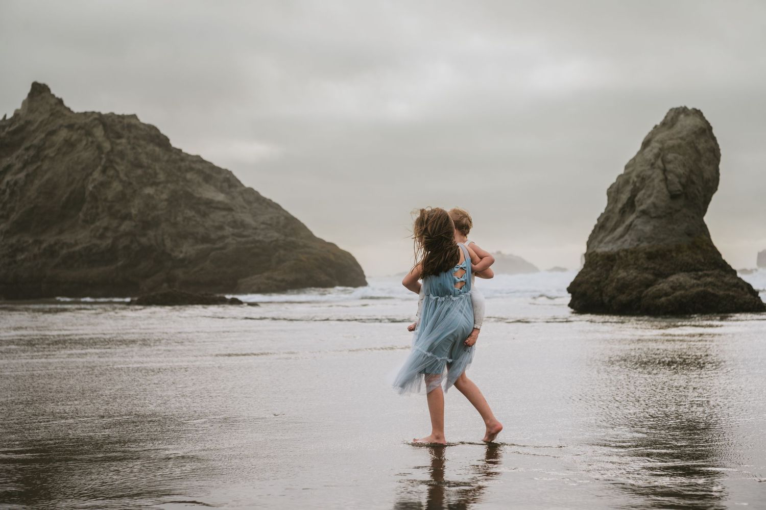 A girl wearing a dusty blue dress, holding a toddler boy walking on the beach.