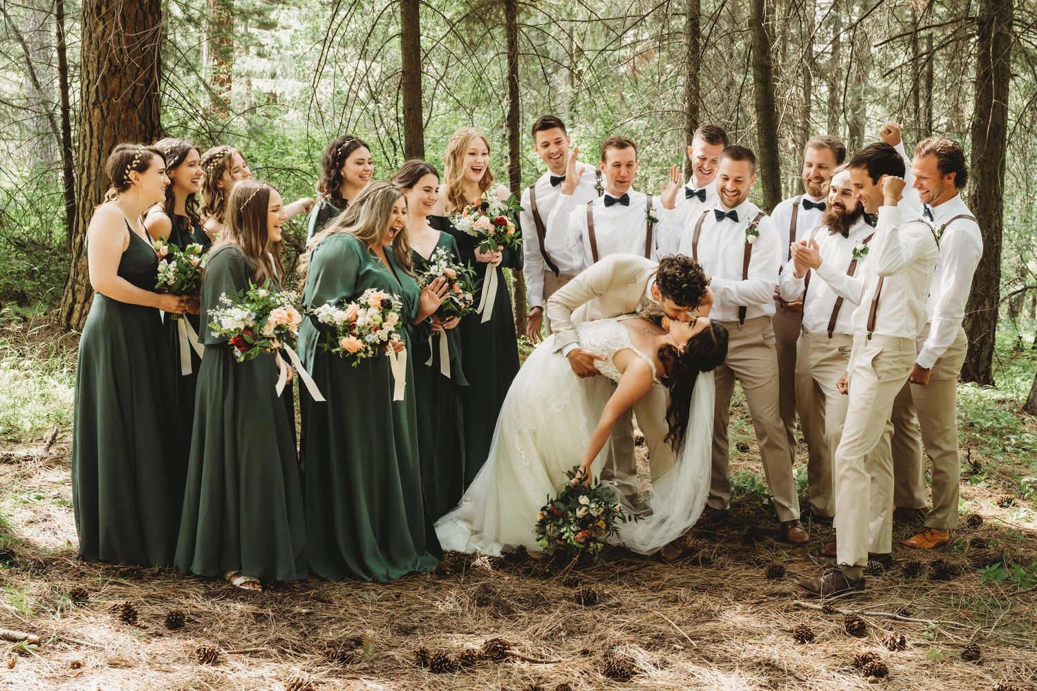 A large wedding party cheering on a bride and groom as they kiss before their wedding ceremony in a forested area near Tri Cities Washington