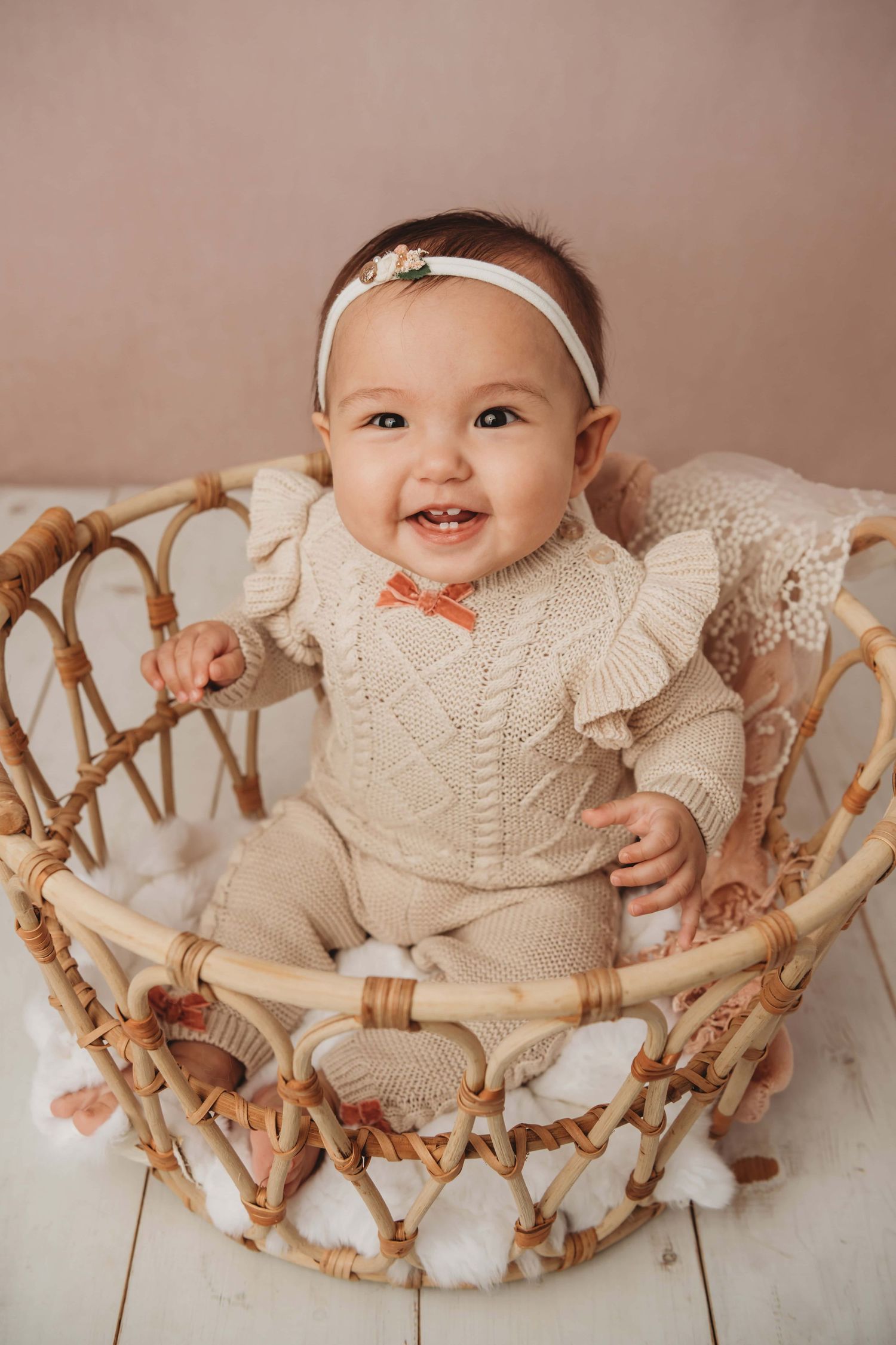 A baby sitting in a brown basket with a pink backdrop and white wooden flooring.