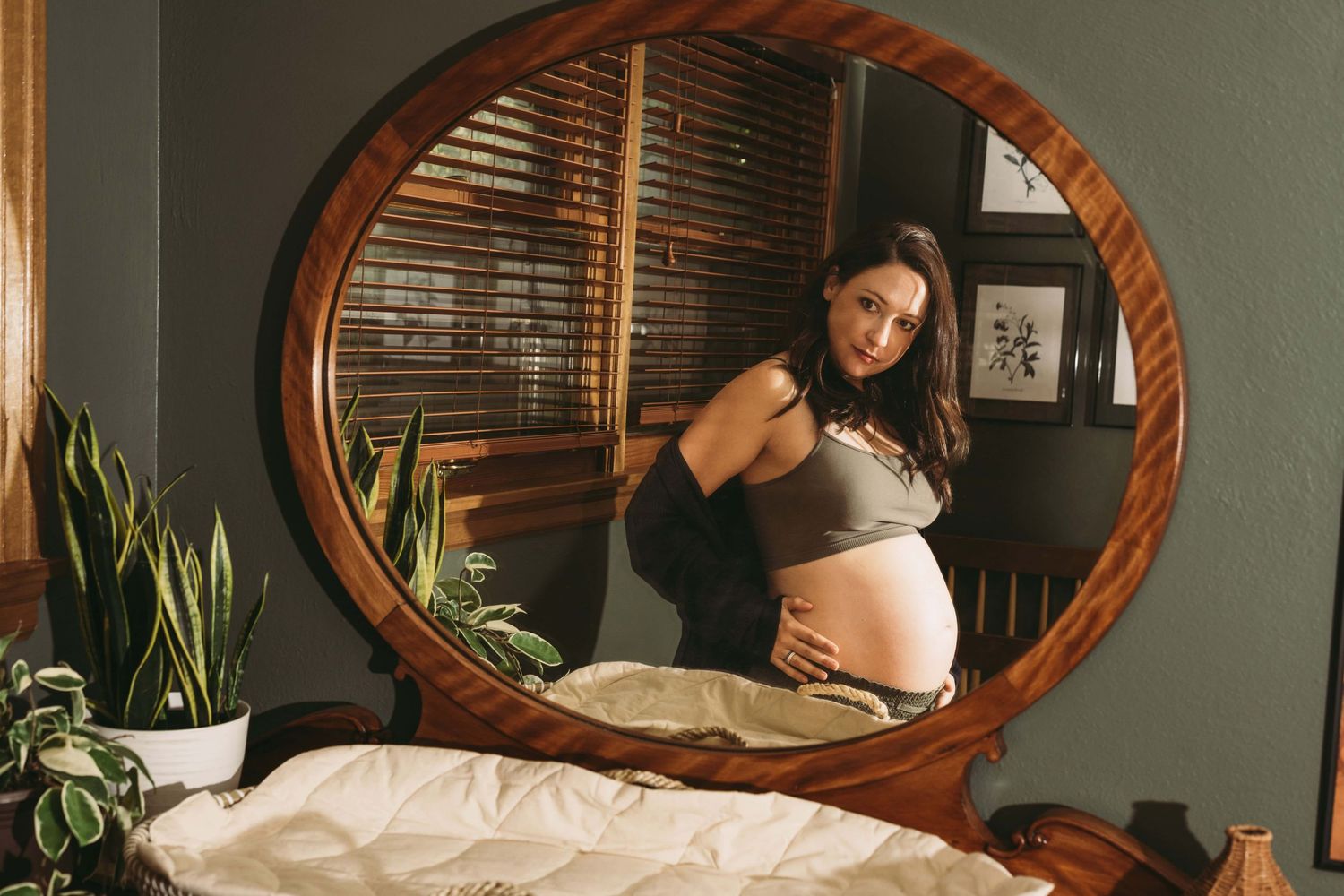 Expectant mother standing in front of a mirror, cradling her baby bump in soft window light during an intimate in-home maternity photoshoot.