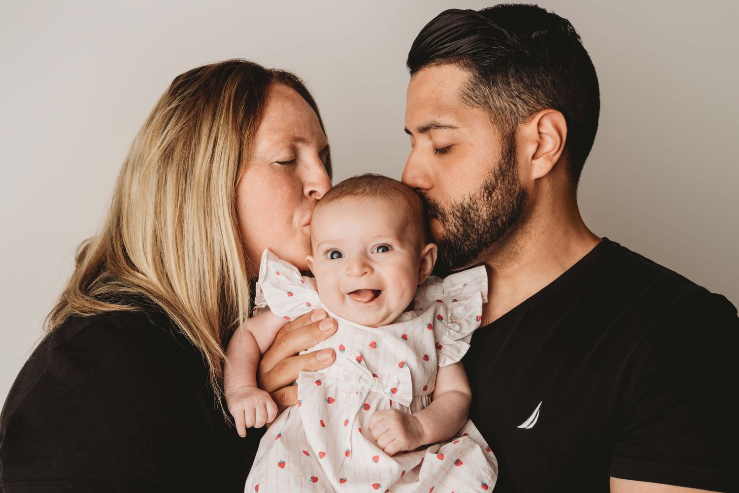 A family portrait of a mom and dad kissing their young baby as she looks at the camera and smiles.