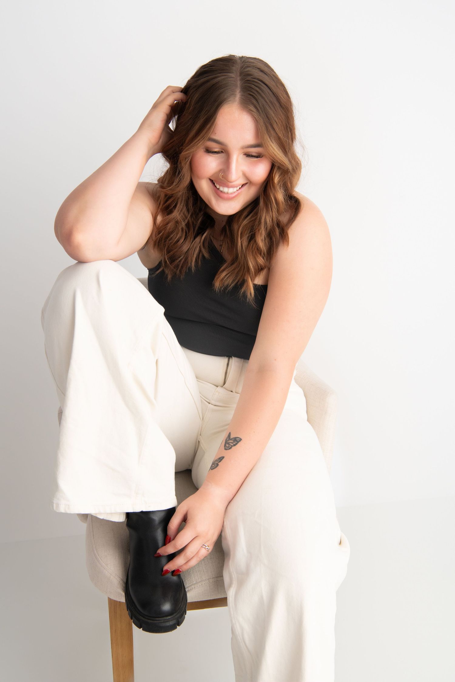 A headshot of a hair stylist sitting on a cream chair looking down and laughing.