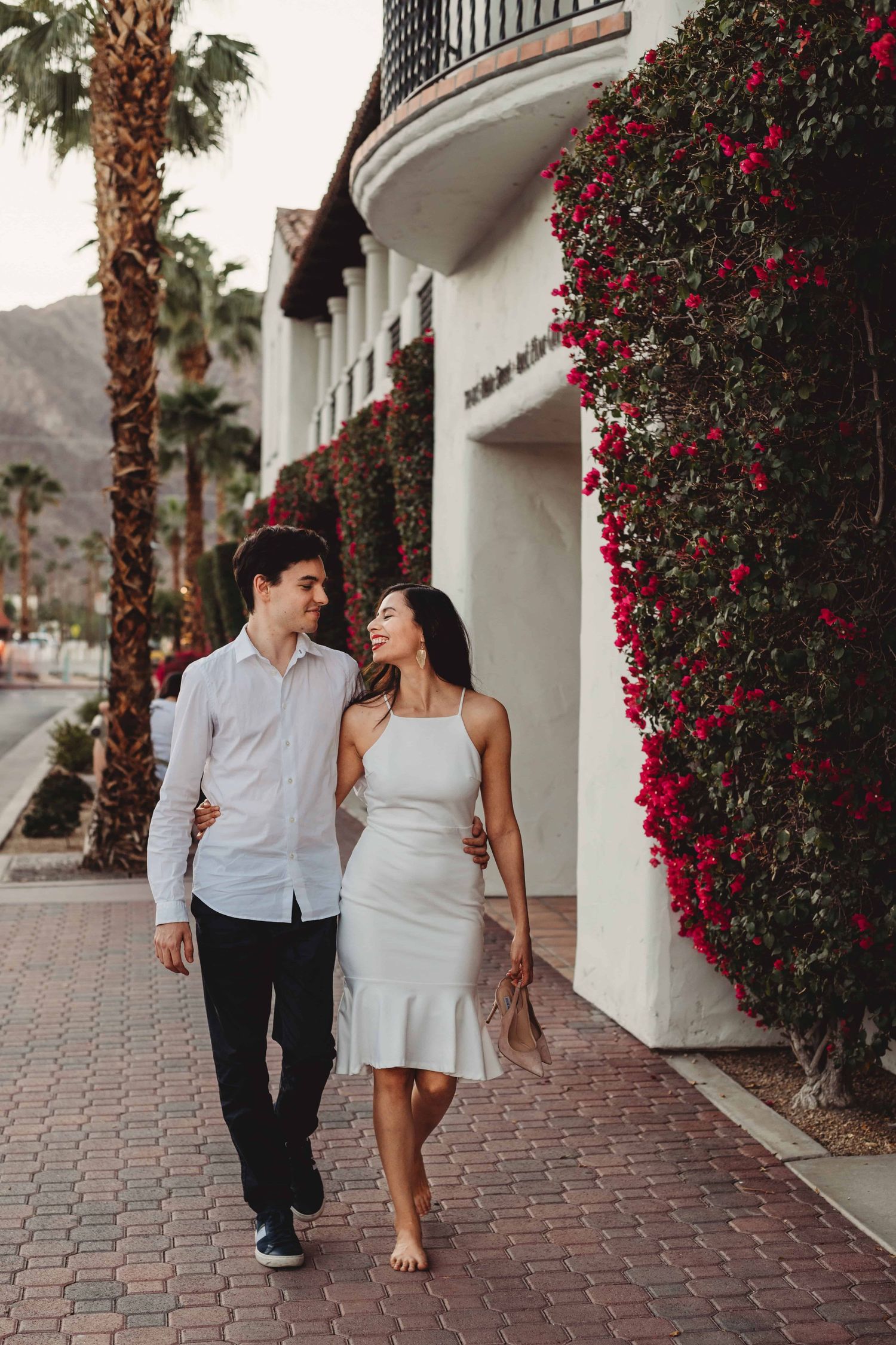 Couple walking arm in arm past a white building with red bougainvillea during an engagement photo session.