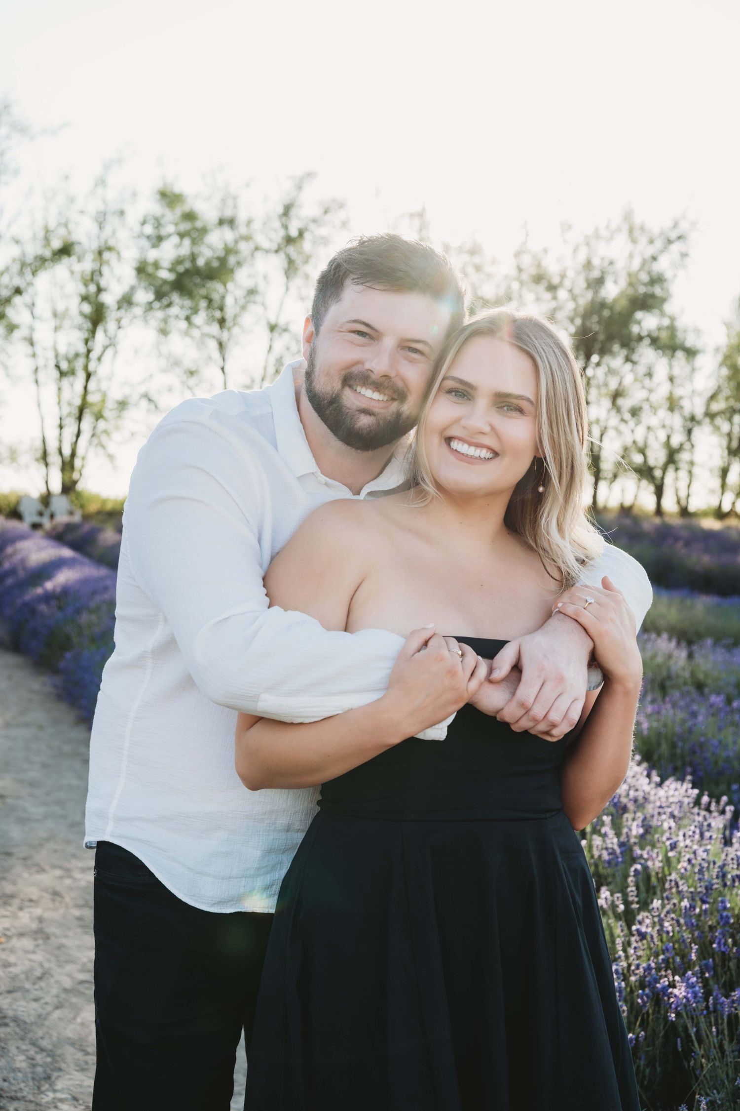 A man hugging his fiancé from behind and both smiling at the camera.