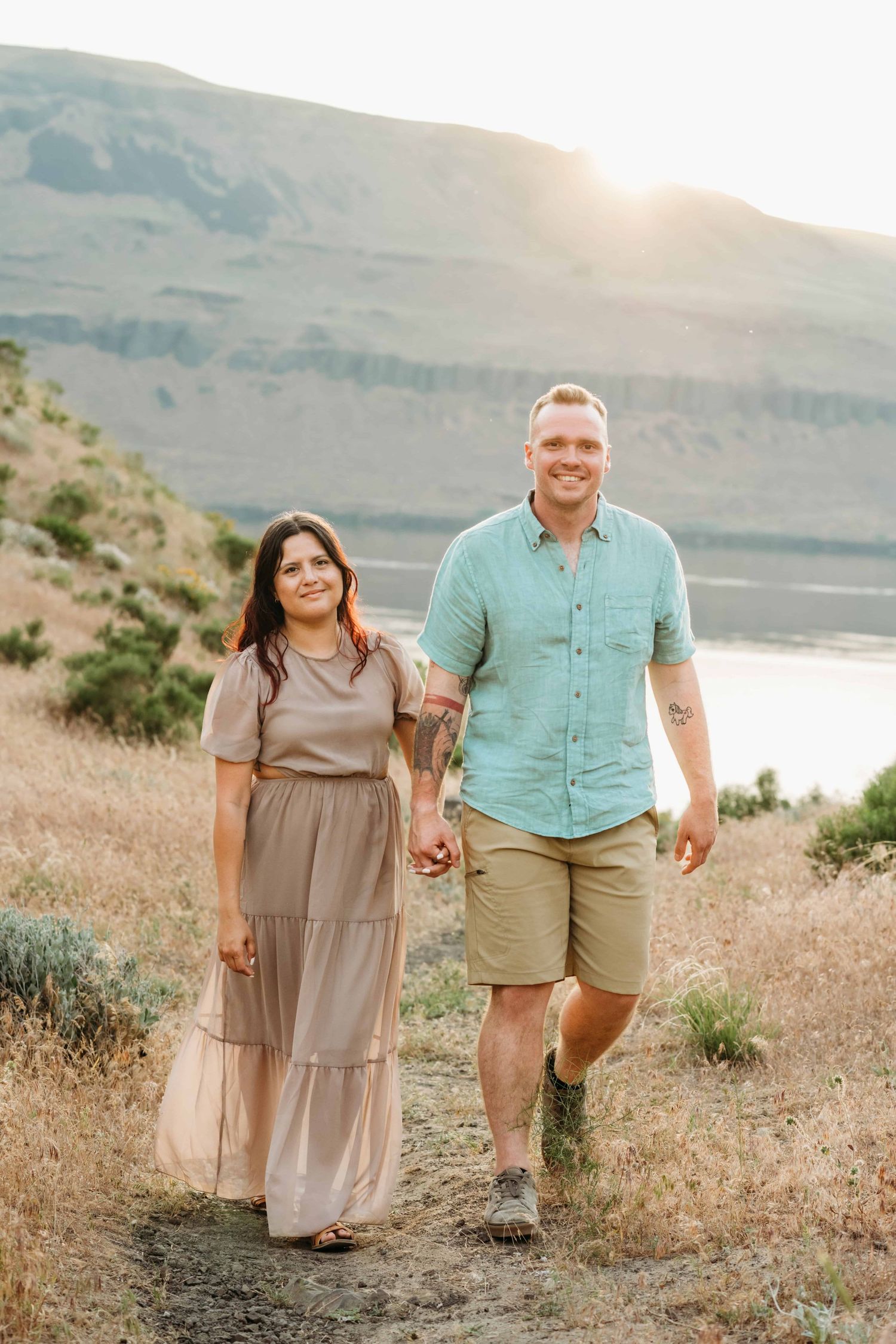A couple walking through a neutral toned field with a river in the background.