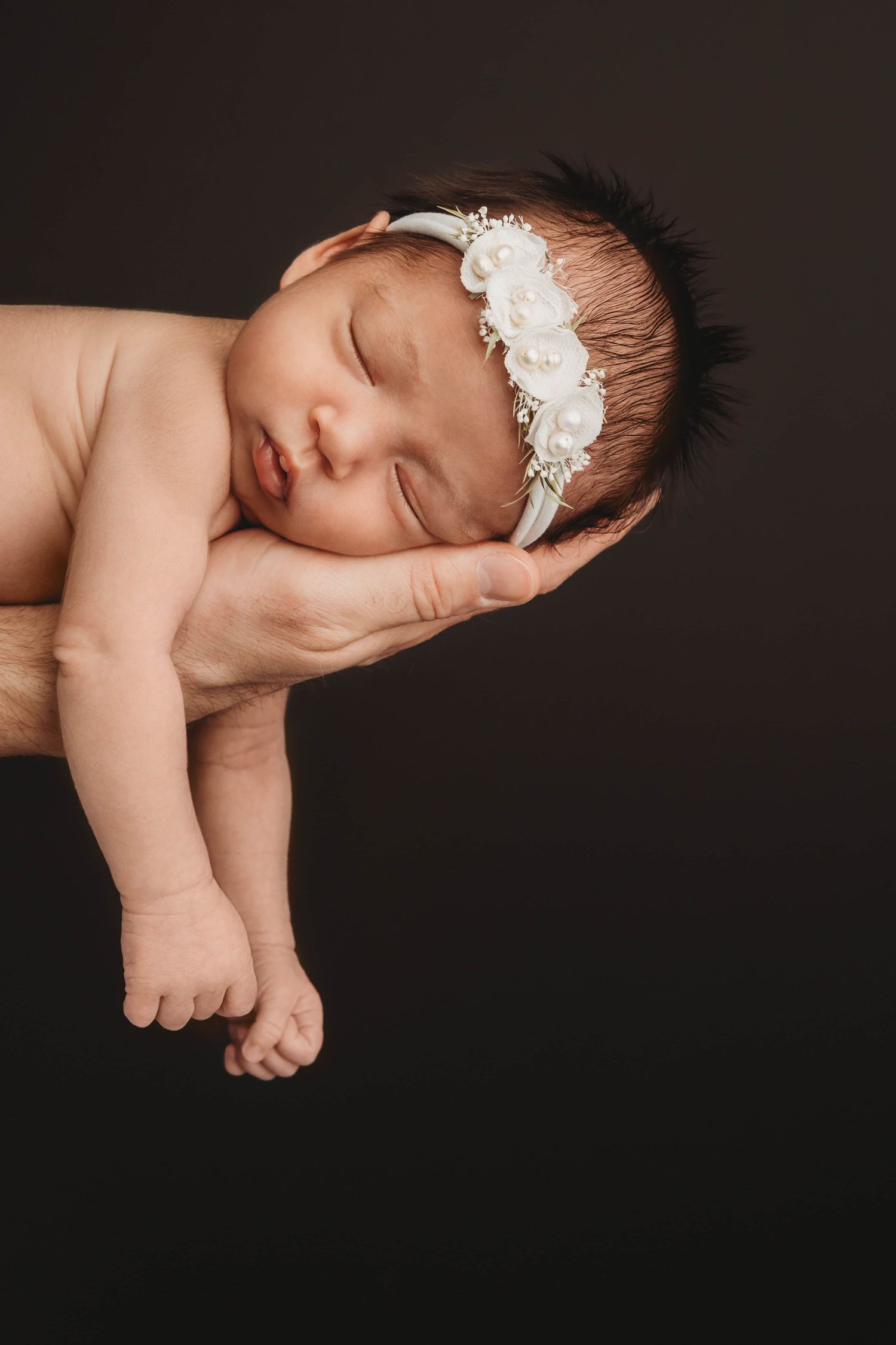 A sleeping newborn wearing a white floral headband sleeping in her father's arm.