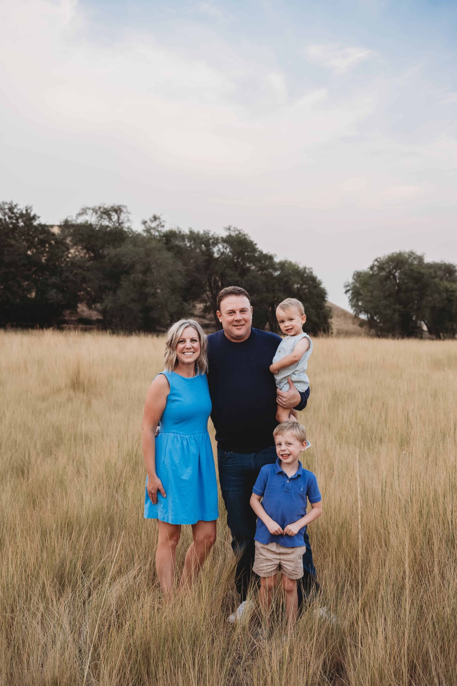 A family with two young boys standing in a field, smiling for the camera during sunset.