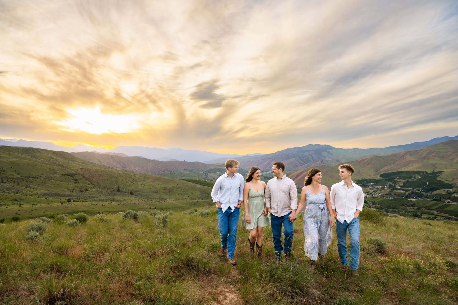 A family of five walking through a grassy field with mountains and a dramatic sunset in the background.