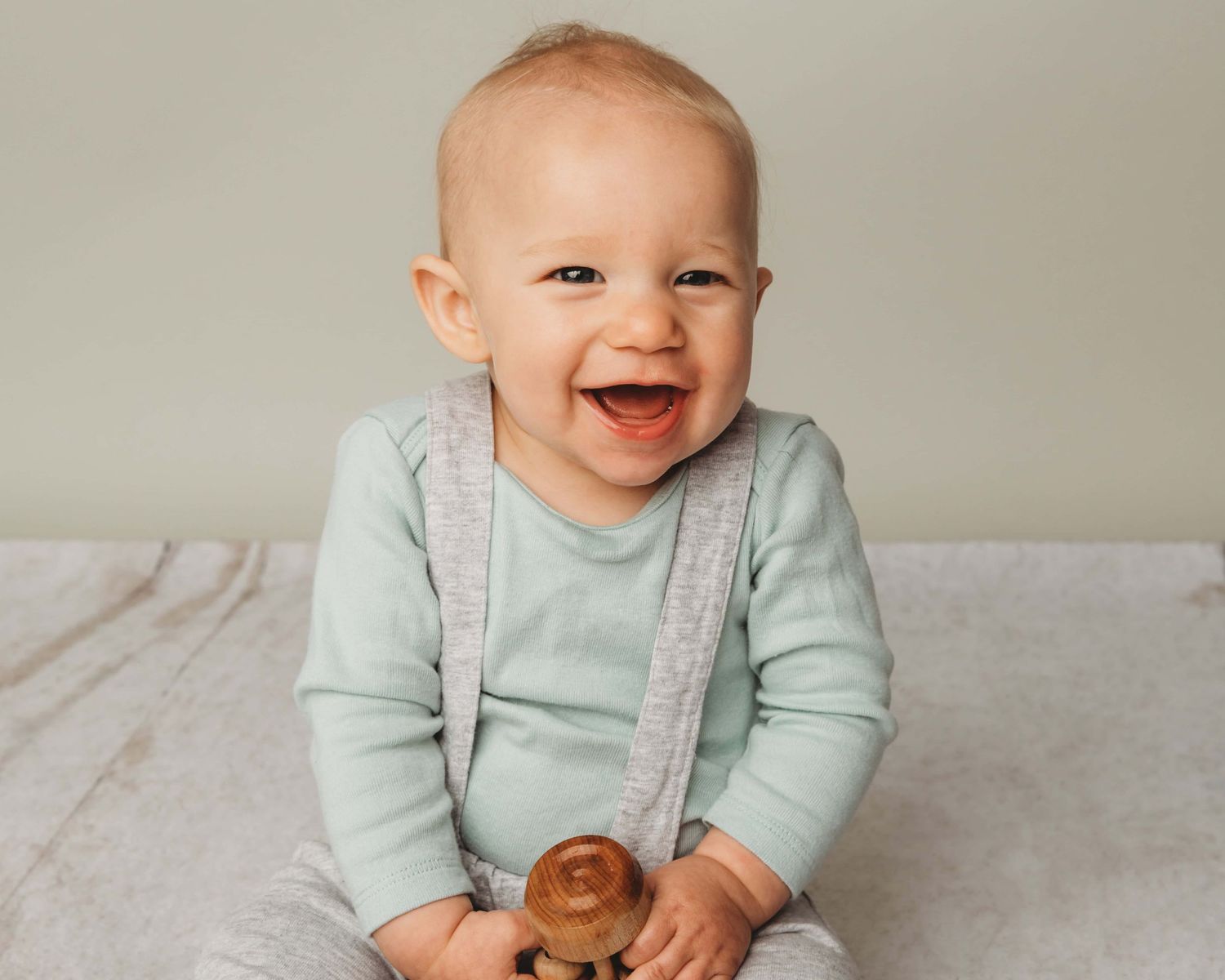 A portrait of a baby wearing gray suspenders and holding a wooden toy while smiling.
