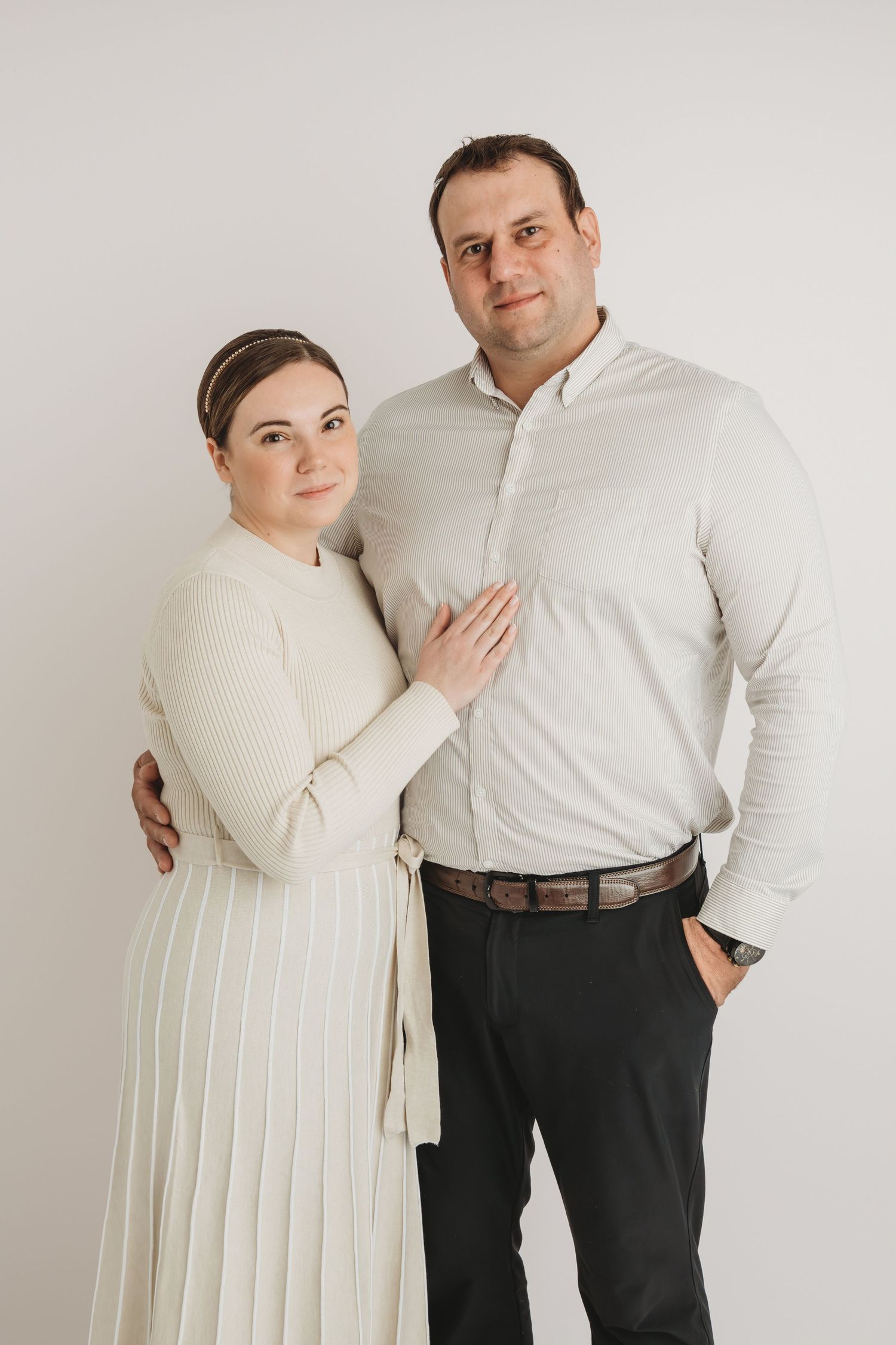 A husband and wife stand on a white studio backdrop smiling for a portrait.