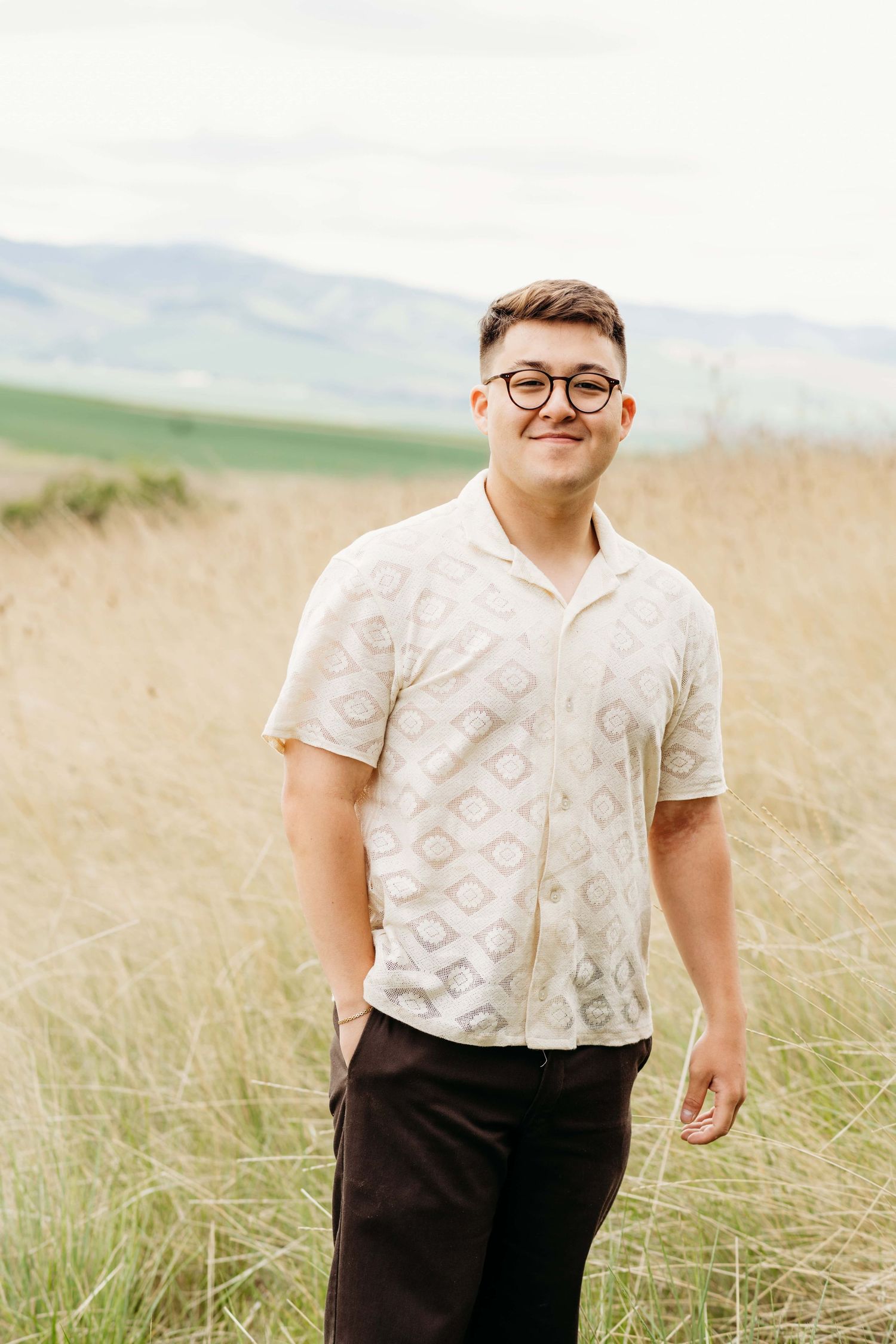 A senior standing in tall grasses with a soft smile on his face and hills in the background.