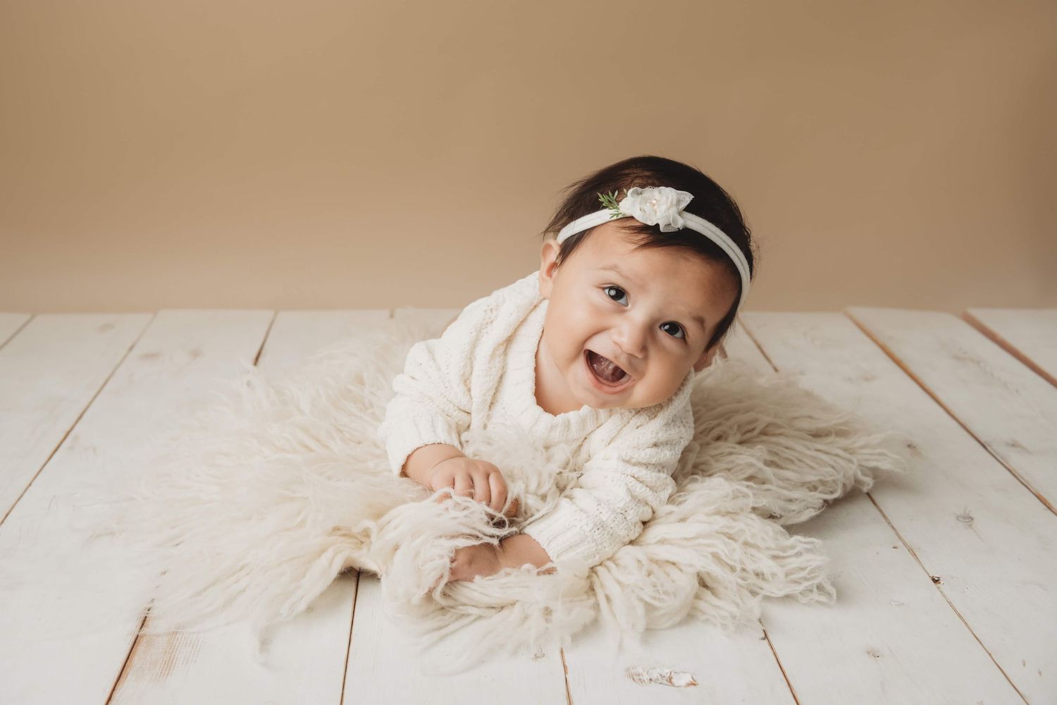 A portrait of a baby wearing all white laying on a soft cream flokati with a brown backdrop.