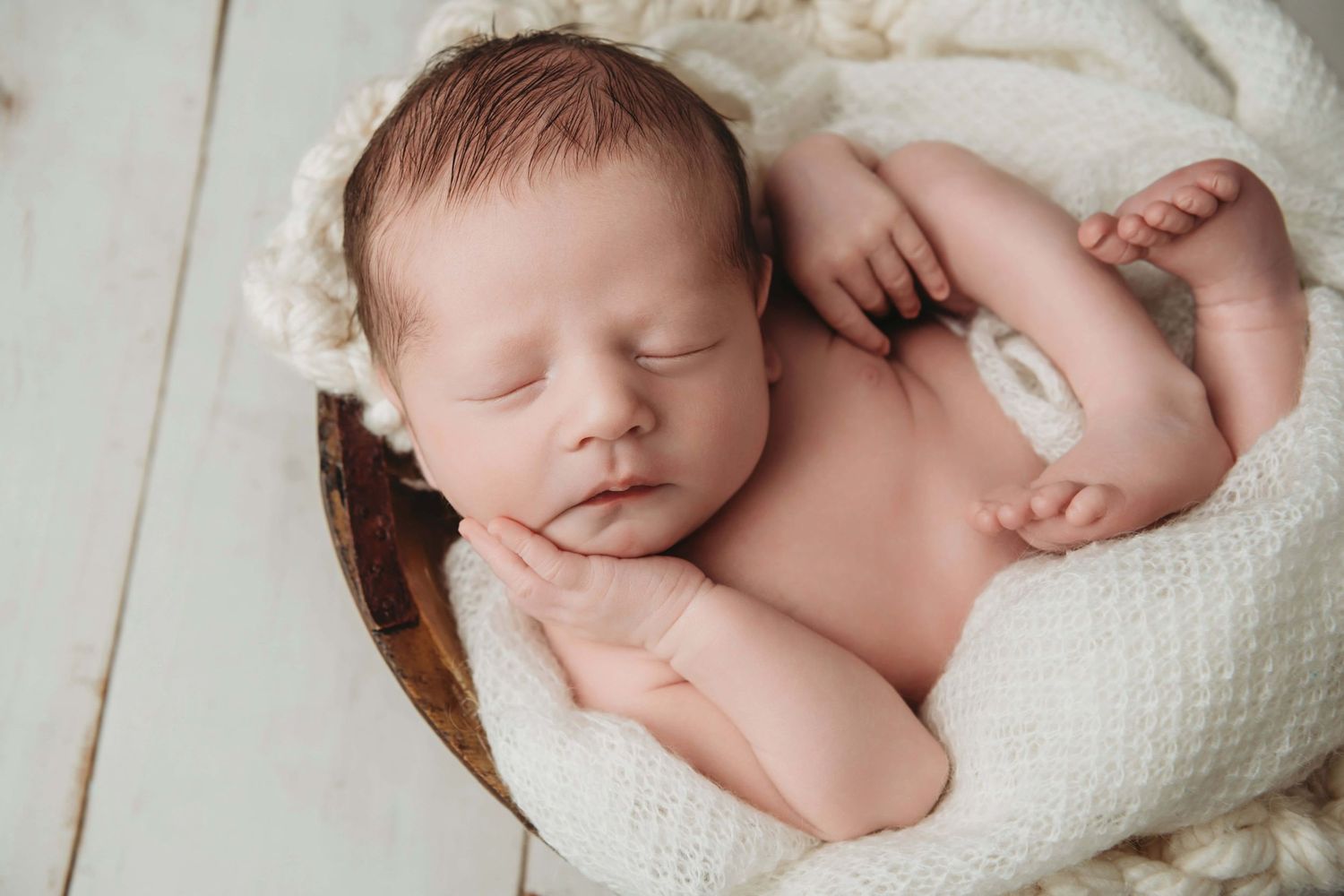 A newborn sleeping in a wooden bowl with cream colored blankets.