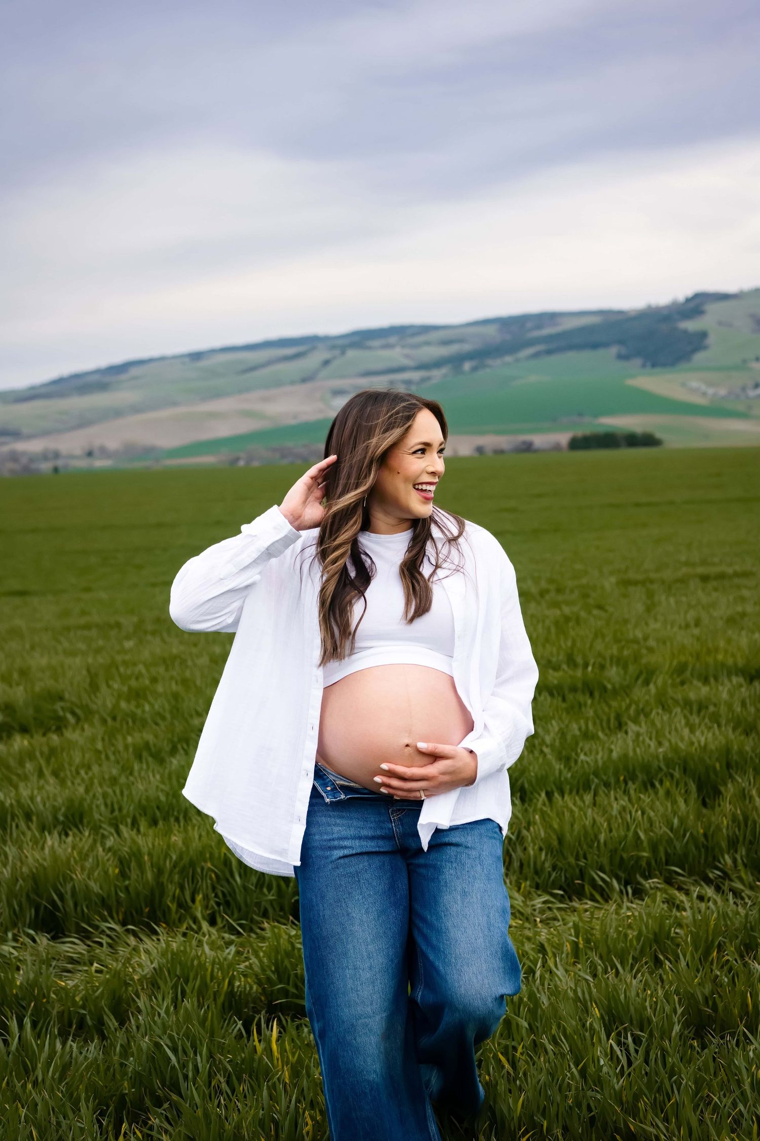 A woman walking through a green grassy field, holding her pregnant belly and looking away from the camera.