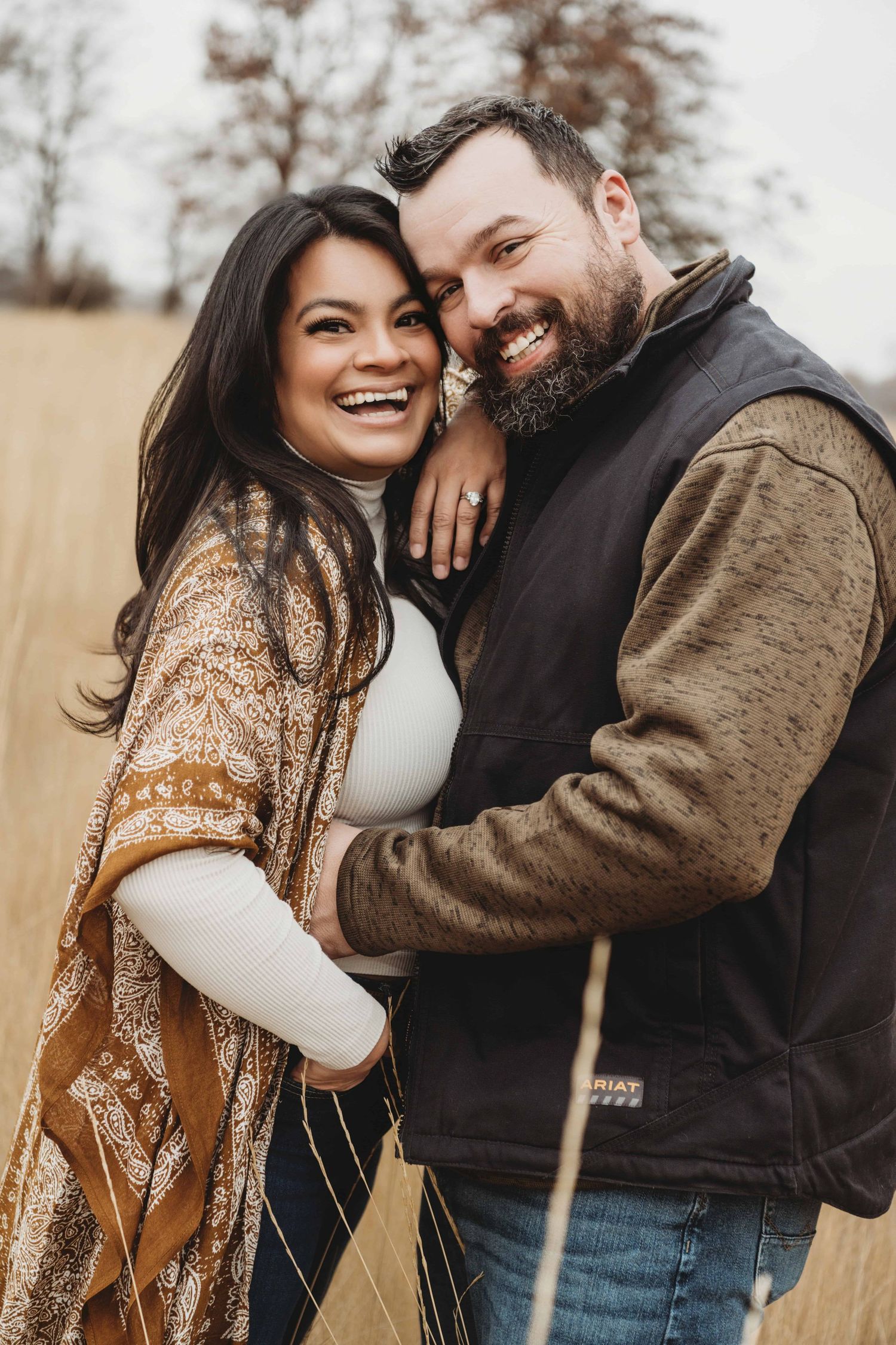A couple wearing neutral brown tones, looking at the camera and laughing.
