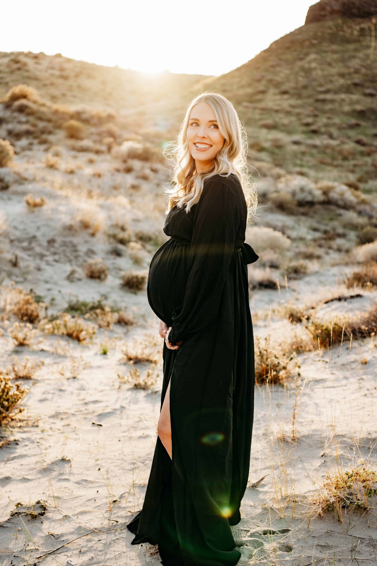 An expectant mother in a black dress stands in a sandy dune with soft sunlight shining in the background.