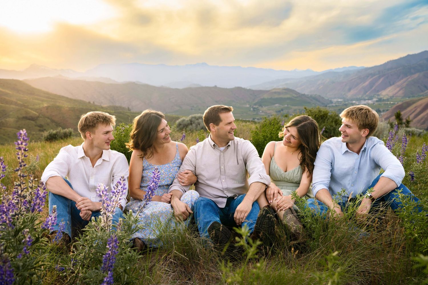 A family sitting in a field with lupin flowers and mountains in the background with a dramatic sunset.