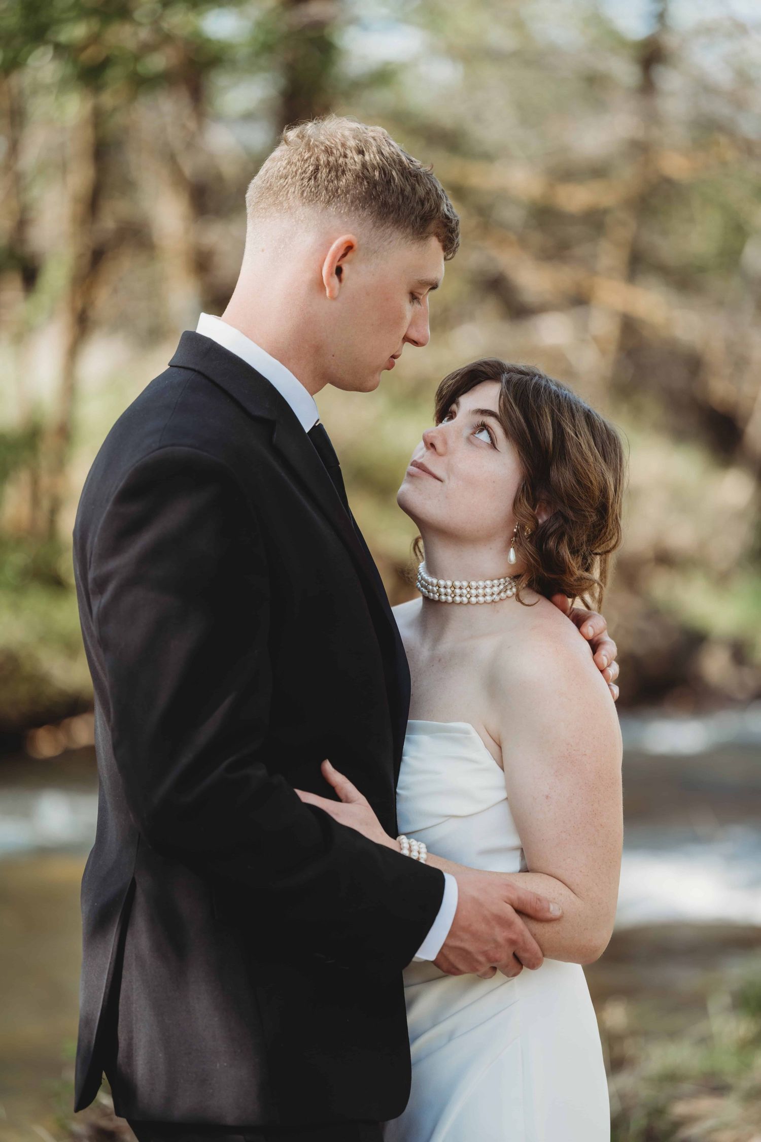 A bride and groom standing in front of a creek and looking at each other with serious expressions on their faces.