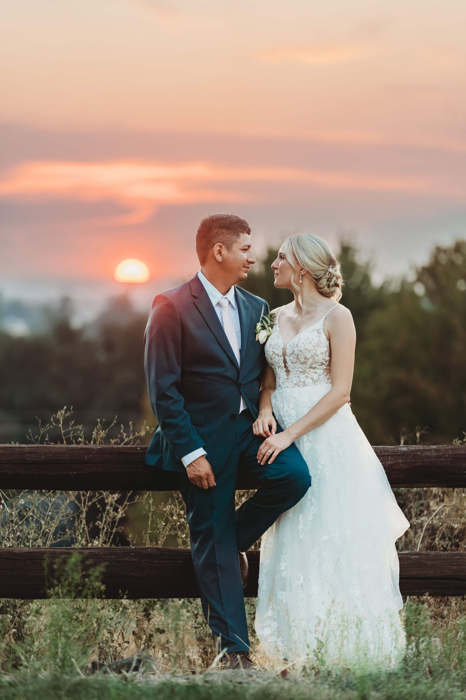 A bride and groom leaning against a wooden fence at sunset and looking at each other.