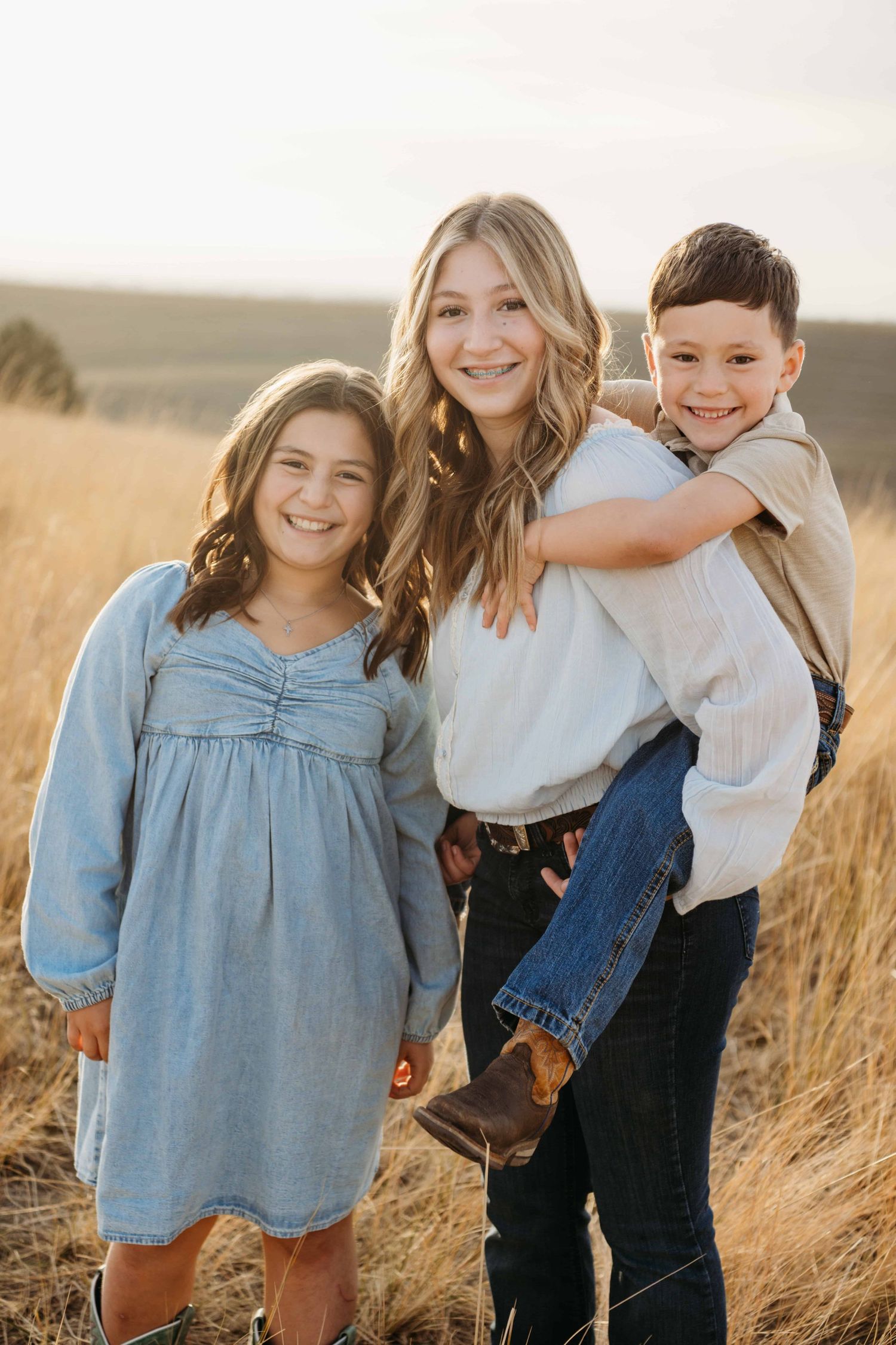 A portrait of three siblings with one on his big sister's back, all smiling at the camera.