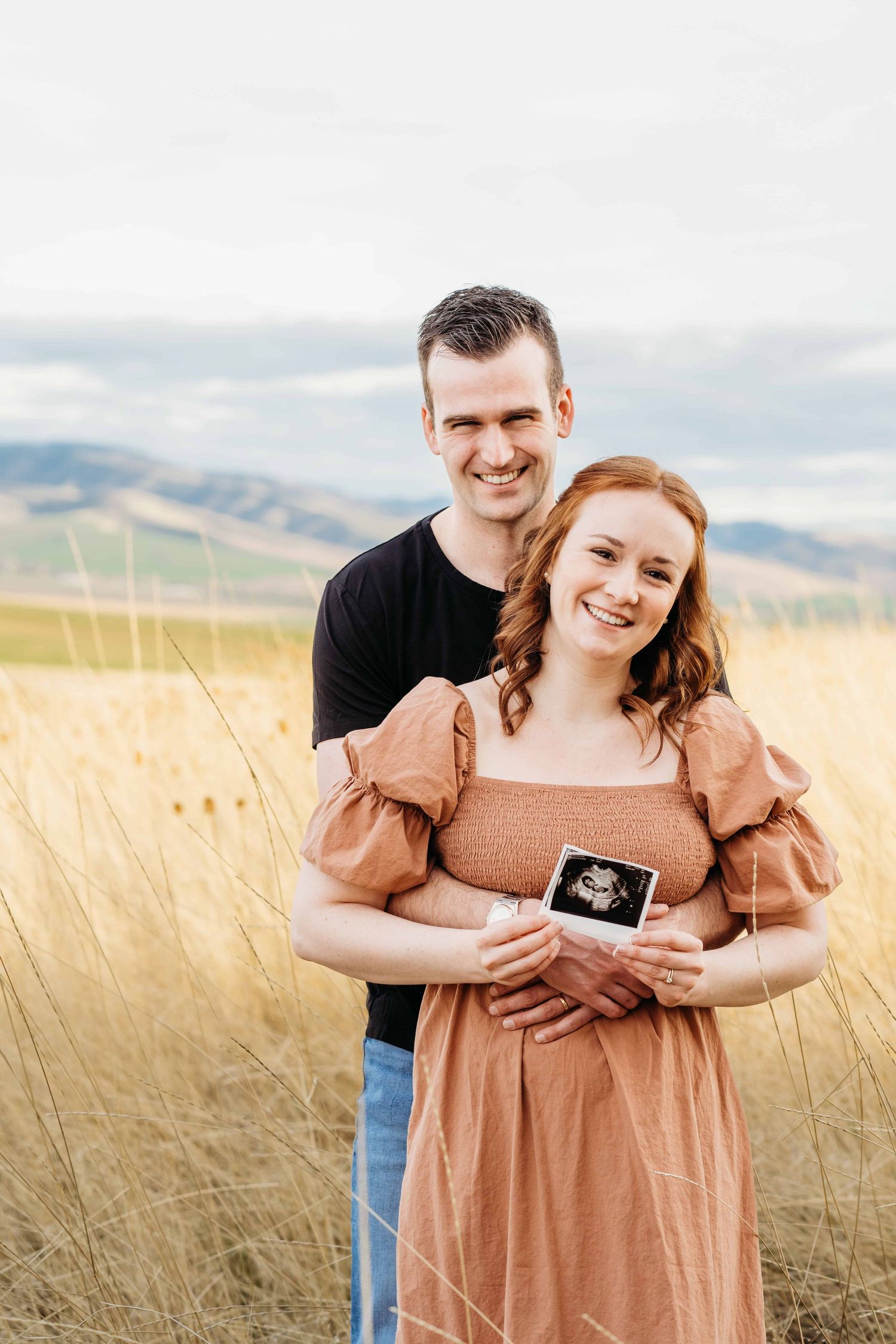 A pregnant woman holding an ultrasound photo while her husband embraces her with rolling hills in the background at Bennington Lake.