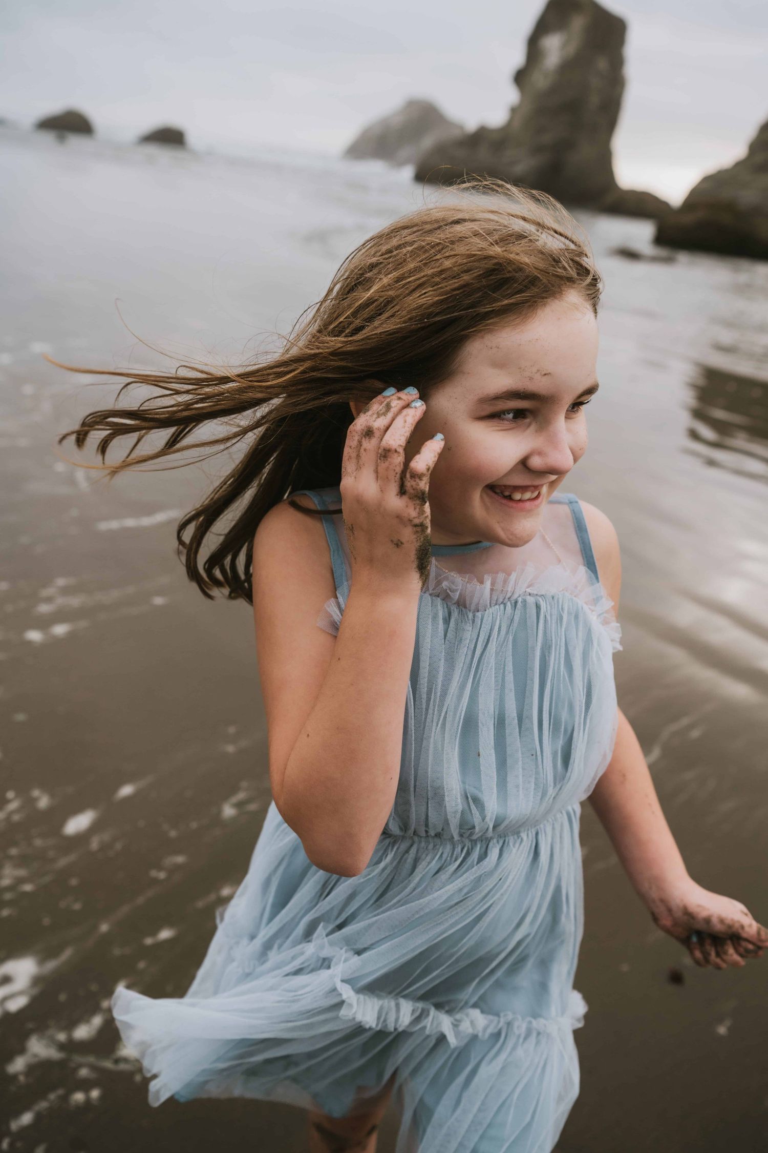 A girl wearing a blue dress standing on the beach with the wind blowing her hair.