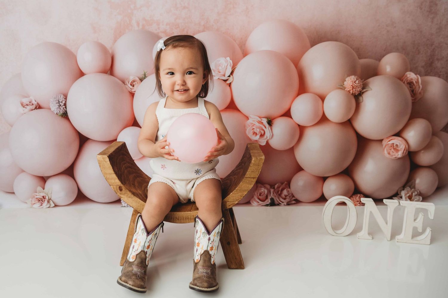 A one year old site on a wooden stool holding a pink balloon.