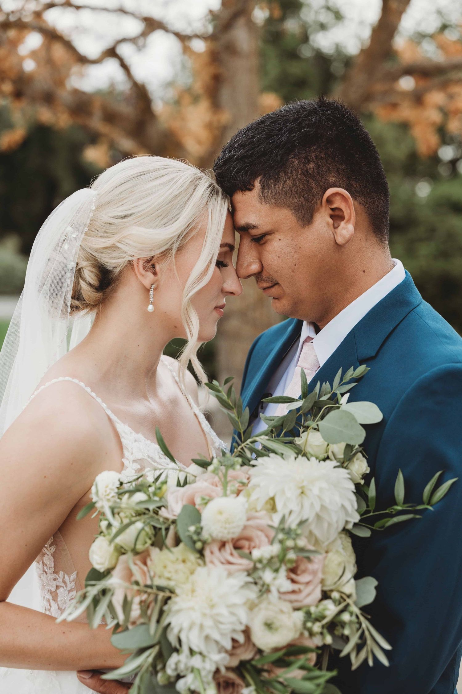 An intimate portrait of a bride and groom with their foreheads touching, with a light bouquet in the foreground.