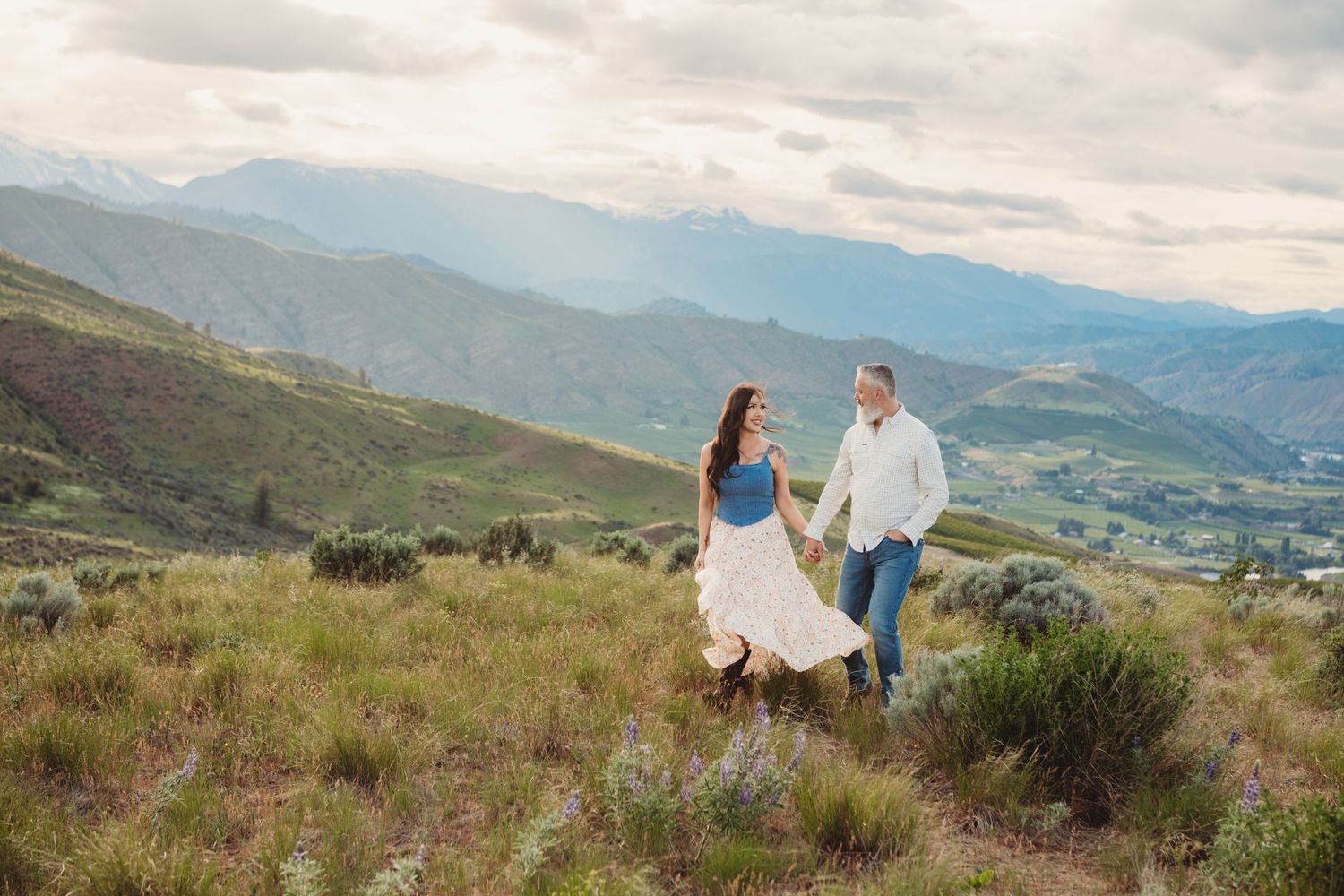 A couple walking through a green grassy field holding hands with mountains and clouds in the background.
