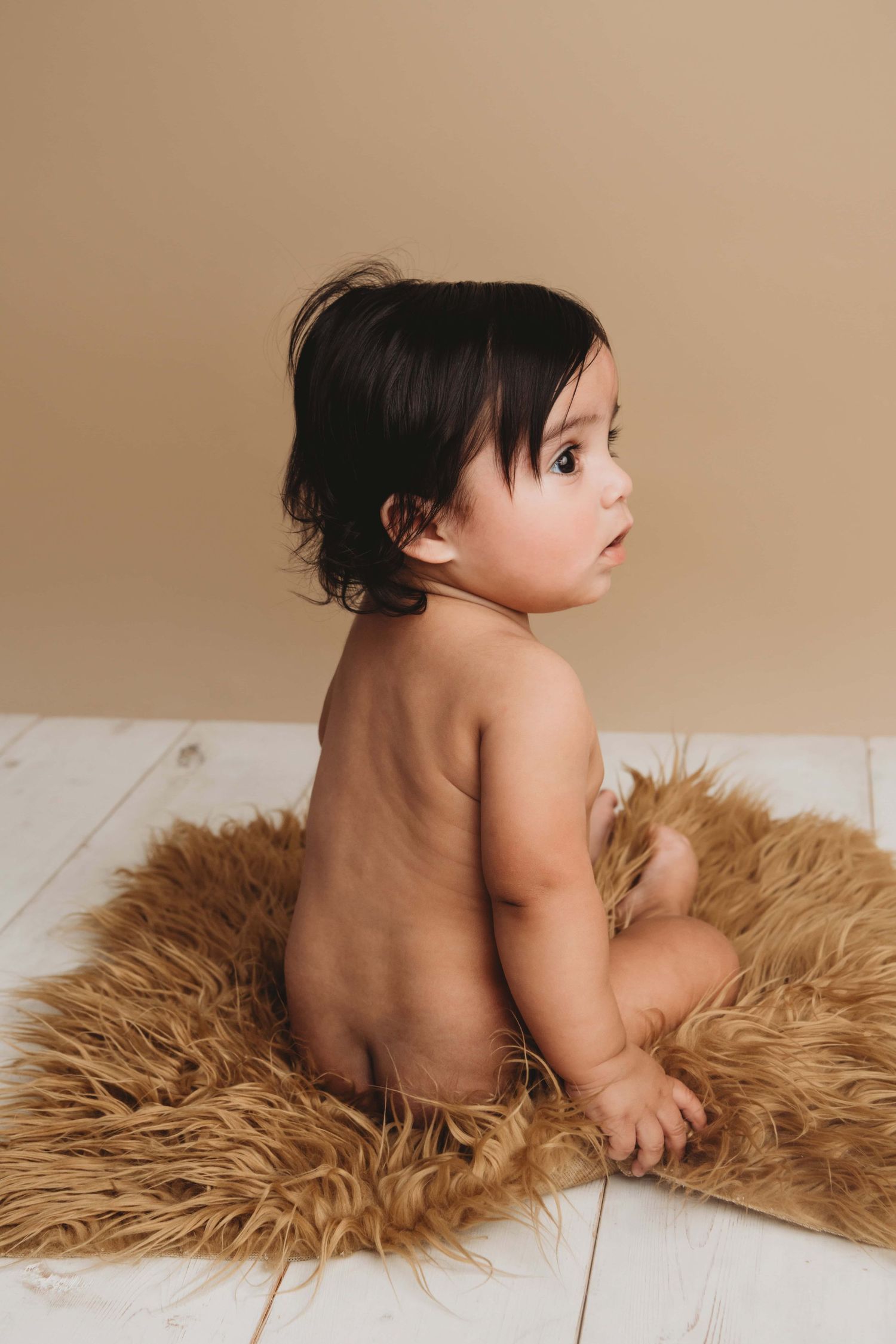 A portrait of a baby with long black hair sitting on a soft brown rug with a relaxed expression.