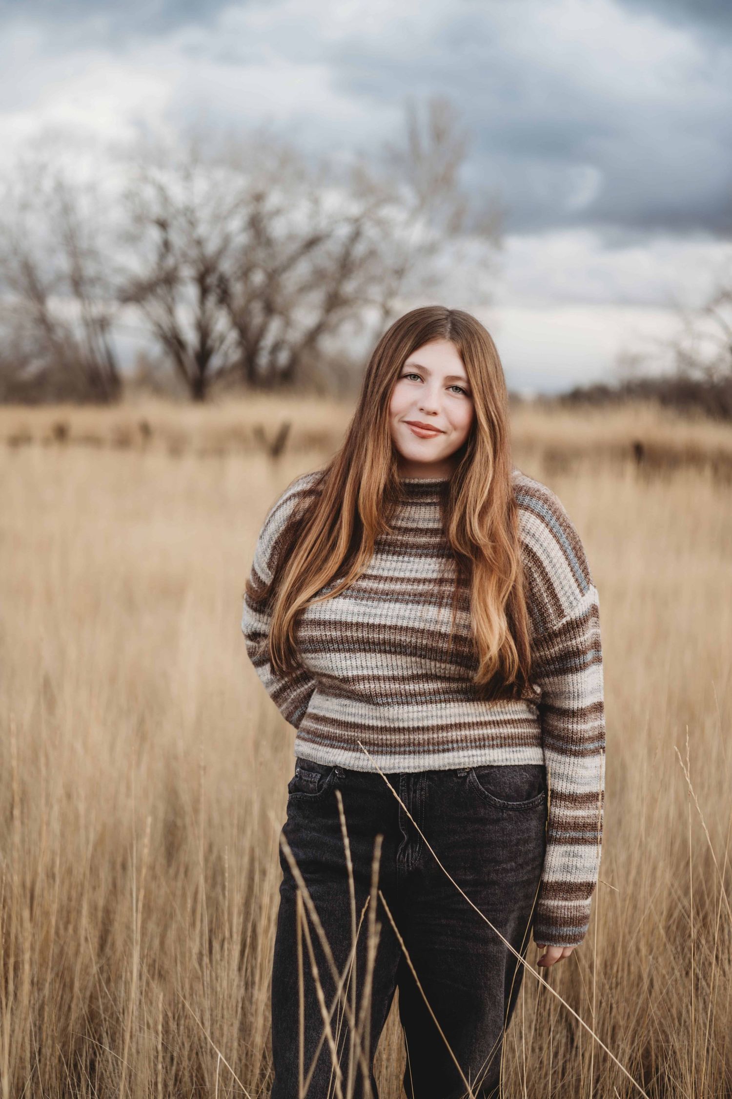 A young woman standing in a field with stormy skies and bare trees during winter in Walla Walla.