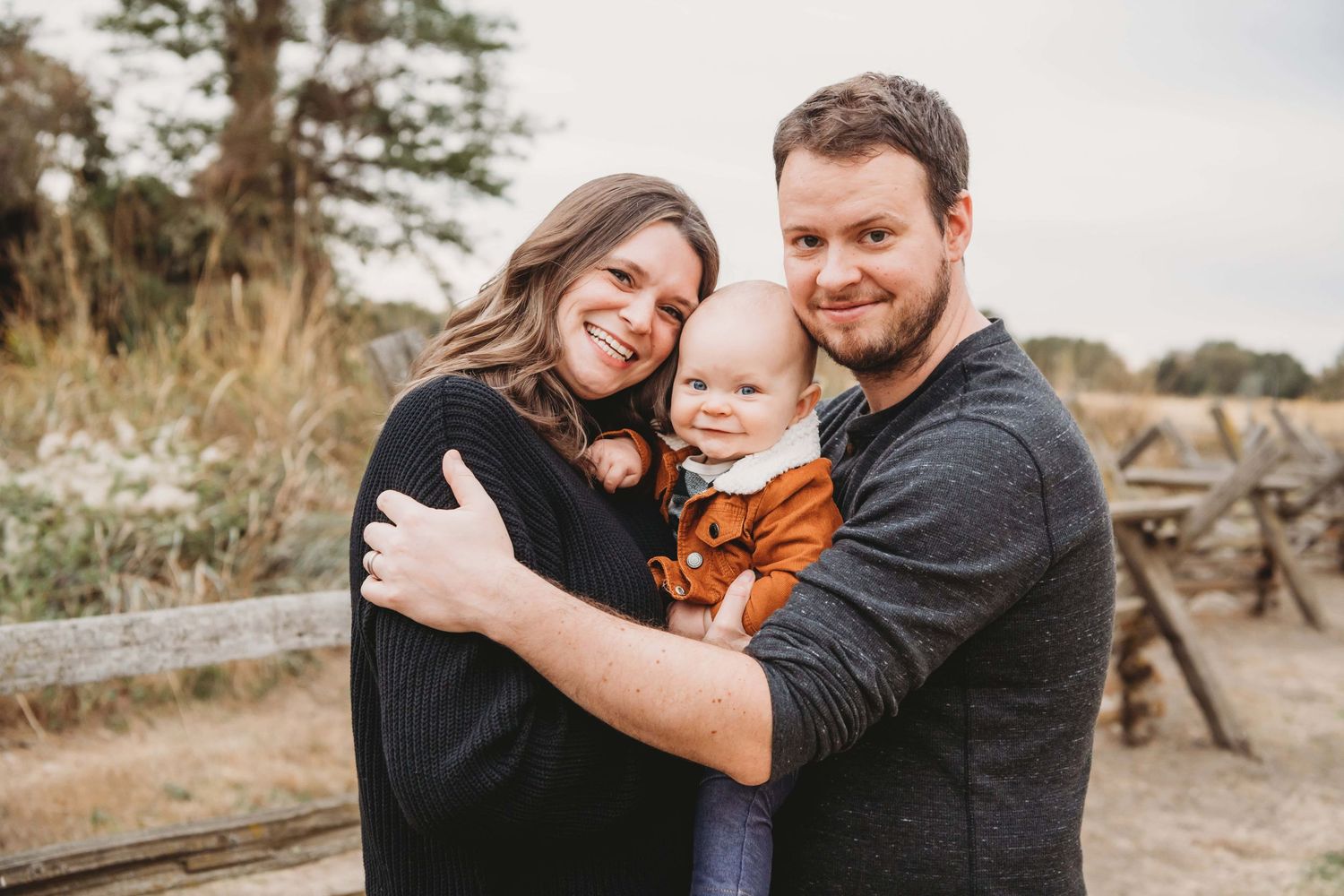 Family portrait by a Walla Walla photographer featuring parents holding their baby outdoors near a rustic fence.
